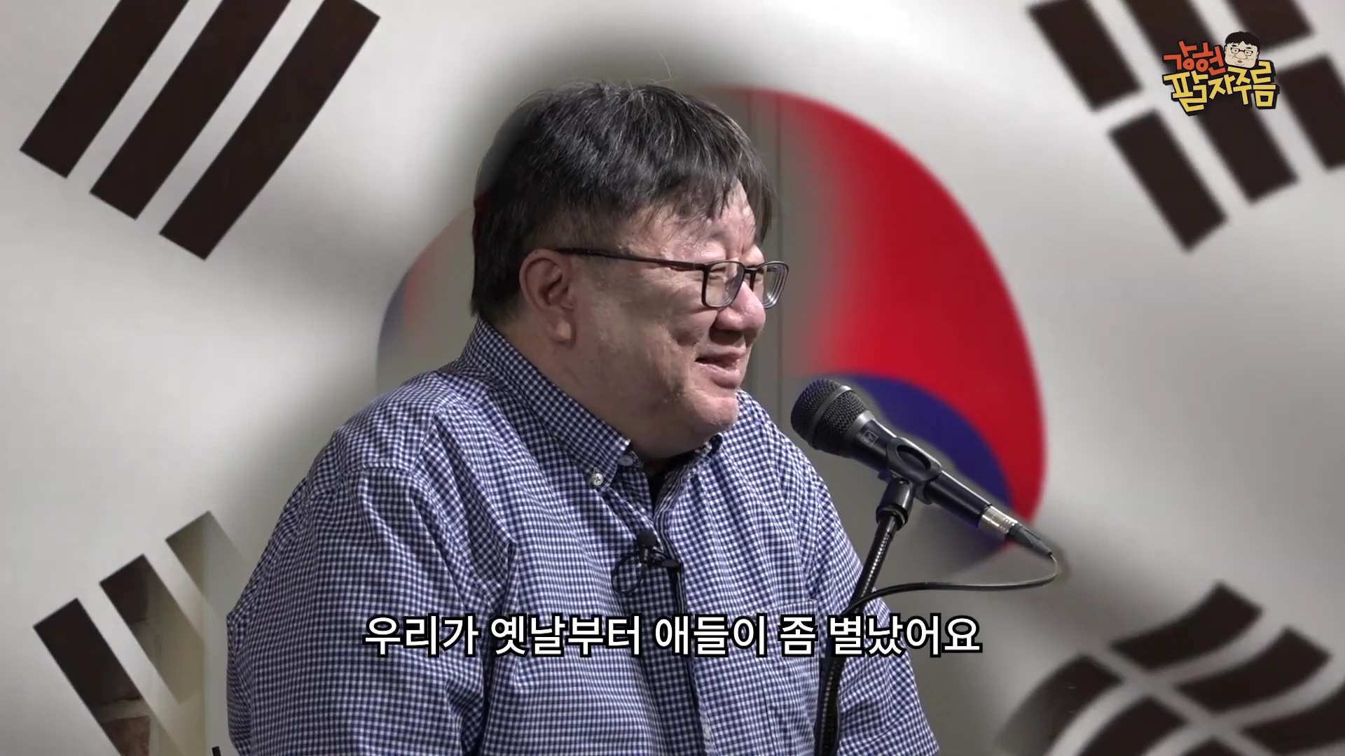 체크무늬 셔츠를 입은 남성이 마이크 앞에서 이야기를 나누고 있으며 뒤편에는 태극기 문양이 흐릿하게 배경으로 보입니다.