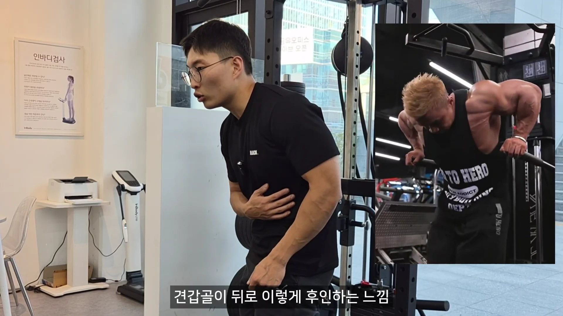 안경을 쓴 운동 지도자가 검은 티셔츠 차림으로 서서 견갑골의 움직임을 설명하고 있으며, 화면 오른쪽 상단에는 보디빌더가 딥스를 수행하는 모습이 담긴 작은 영상이 겹쳐져 있다.