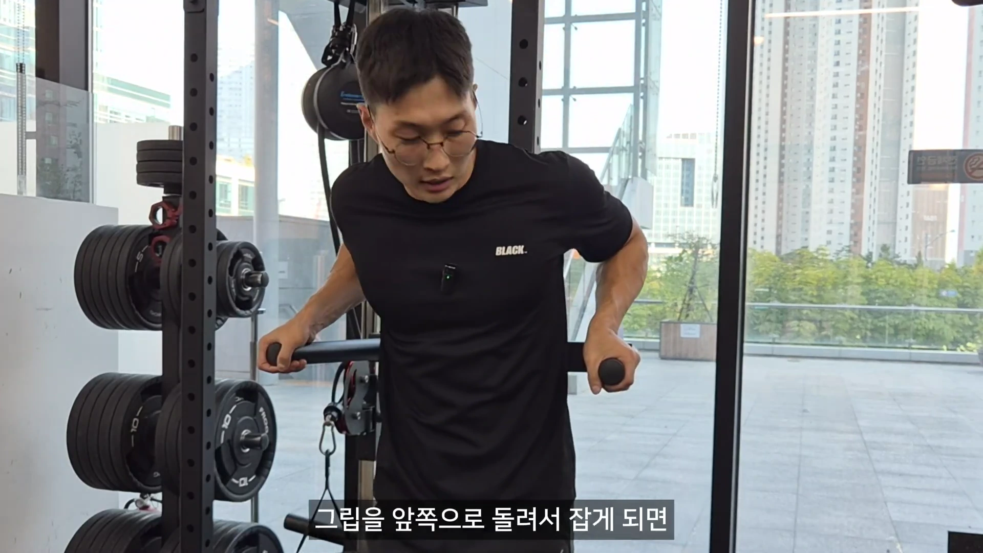 실내 체육관에서 검은색 티셔츠를 입은 남성이 딥스 머신의 그립을 잡고 자세를 취하고 있으며, 하단에는 자막이 삽입되어 있다.
