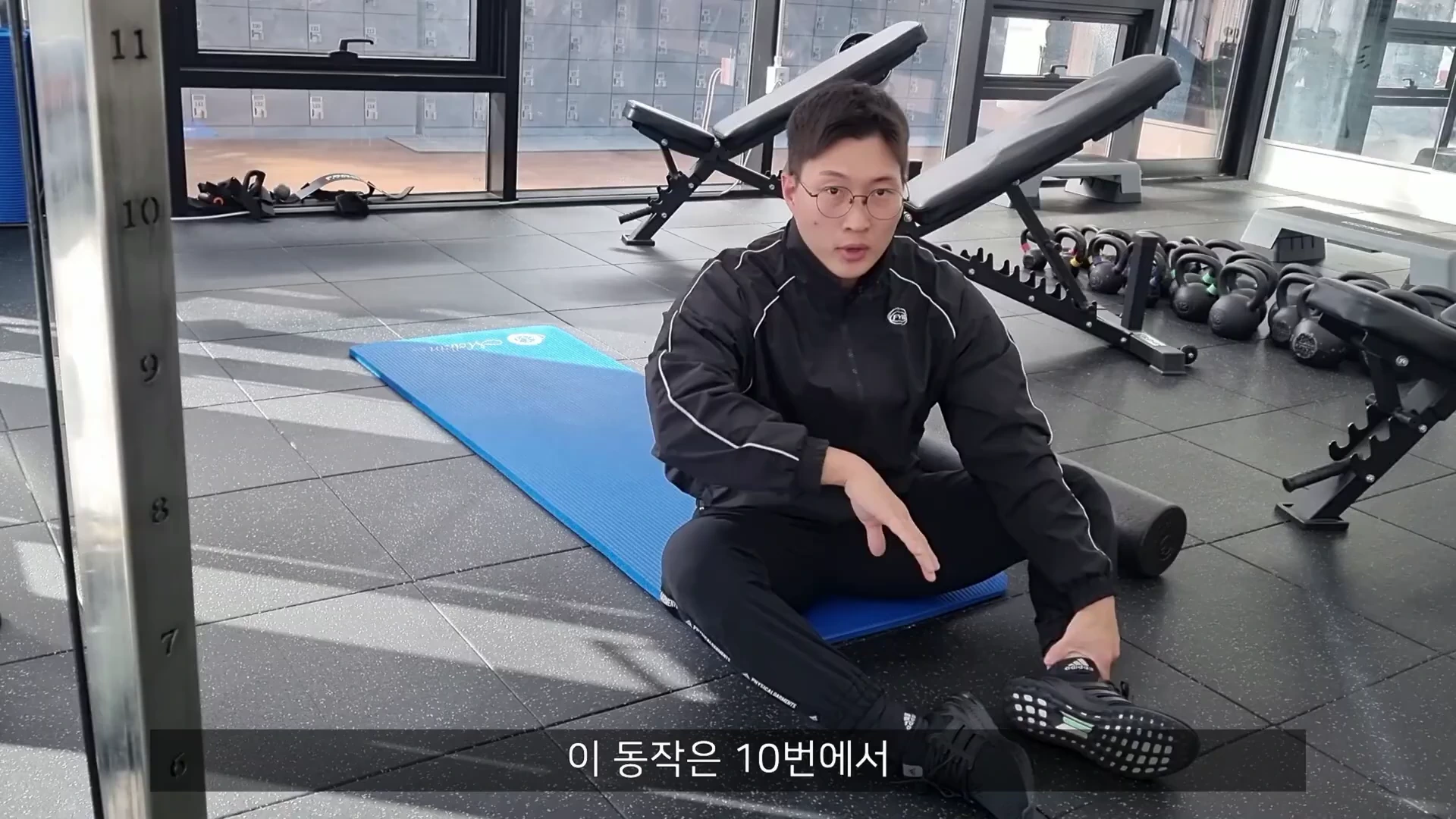 실내 체육관 바닥에 깔린 파란색 요가 매트 위에 안경을 쓴 남성이 앉아 동작을 설명하고 있다.