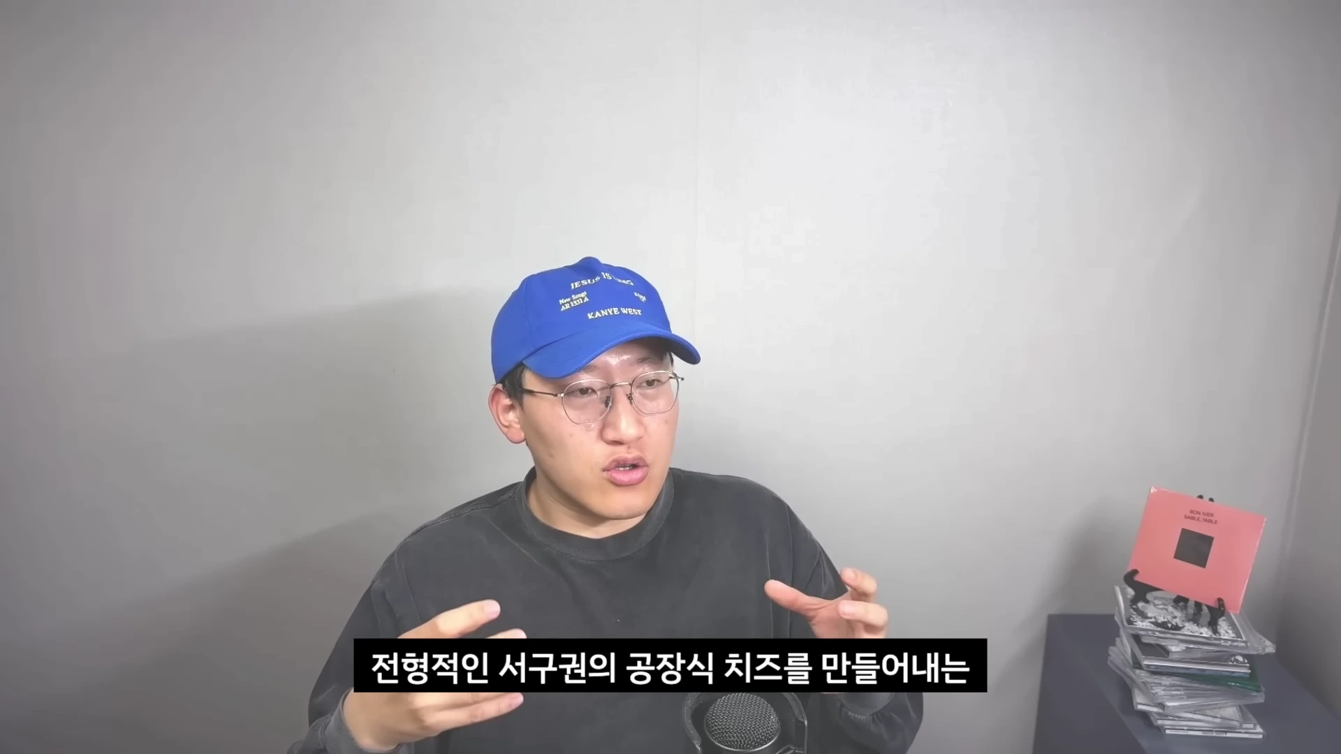 A man wearing a blue cap and glasses talks in front of a plain background while gesturing with his hands.