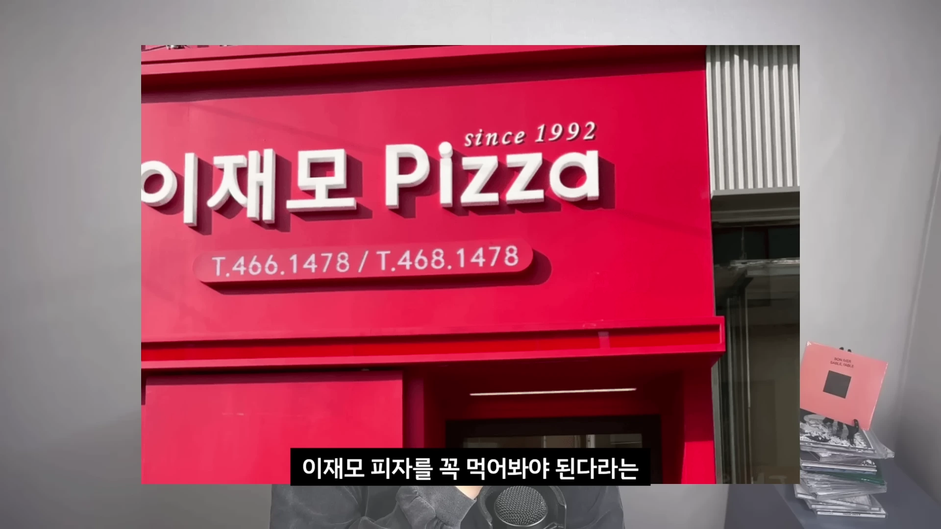 since 1992라는 문구가 적힌 빨간색 이재모피자 간판 아래로 한 사람이 마이크 앞에서 이야기하고 있는 모습