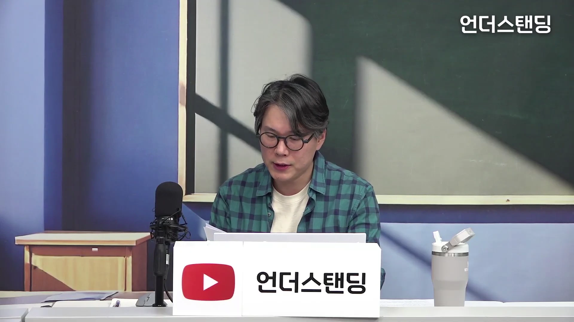 안경을 쓴 남성이 체크무늬 셔츠를 입고 책상 앞에 앉아 자료를 살펴보고 있는 스튜디오 인터뷰 장면