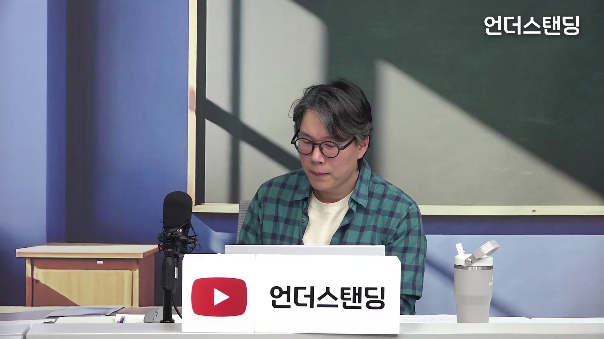 안경을 쓴 남성이 체크무늬 셔츠를 입고 책상에 앉아 마이크 앞에 있는 모습