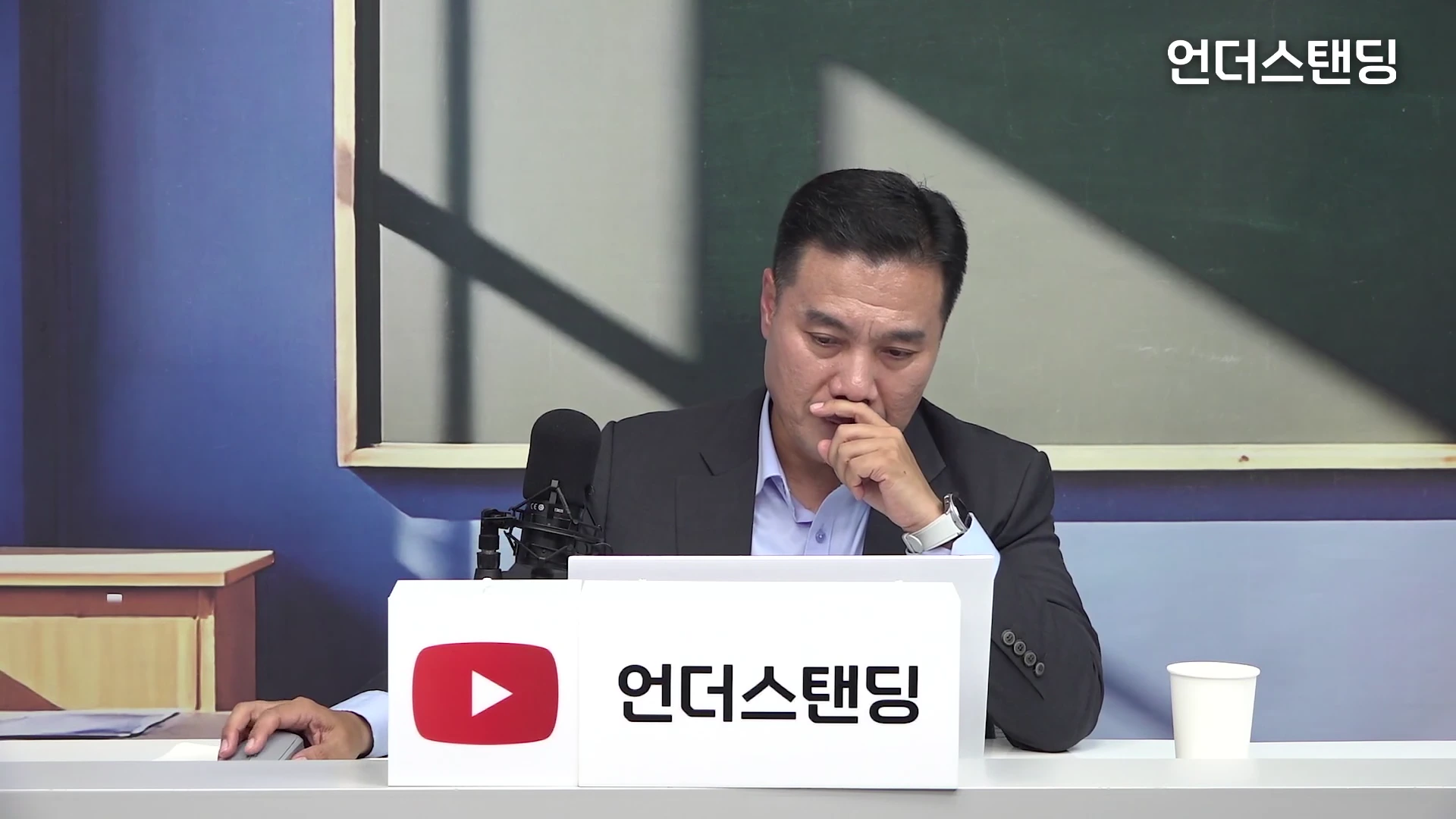 남성이 정장 차림으로 스튜디오 책상 앞에 앉아 한 손으로 입을 가린 채 생각에 잠긴 모습