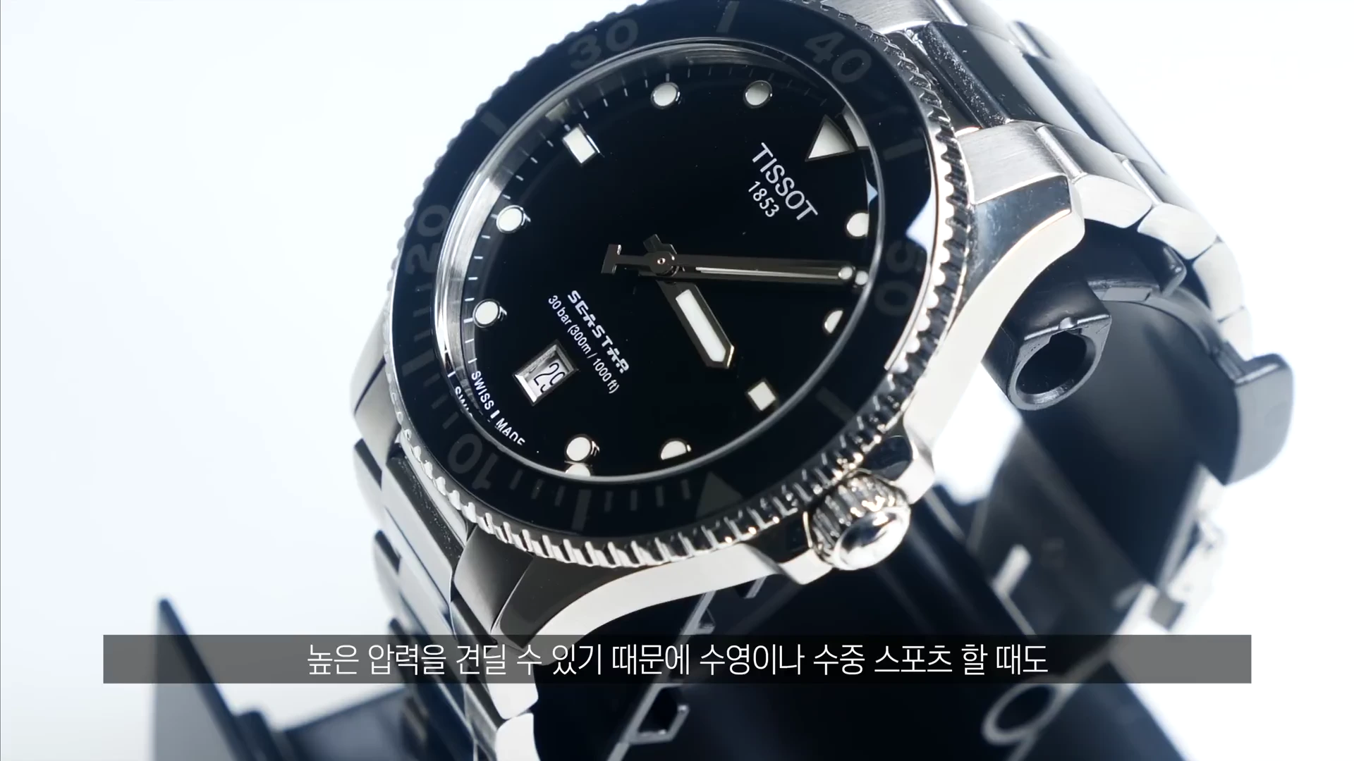 검은색 다이얼과 베젤을 갖춘 티쏘 다이버 시계가 스탠드에 거치되어 있으며 하단에는 자막이 포함되어 있습니다.