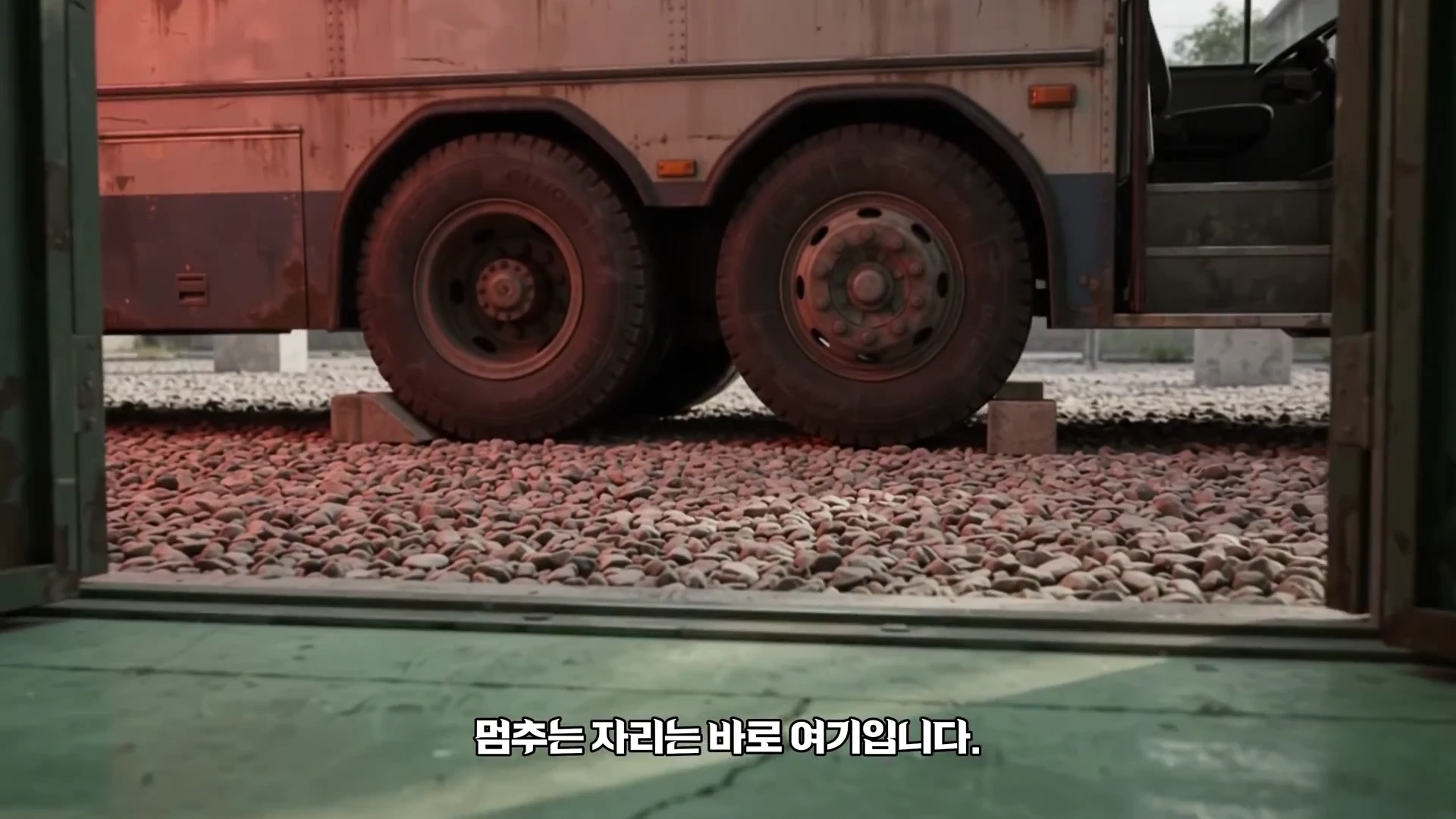 자갈밭 위에 놓인 버스의 뒷바퀴와 차체 하단부 모습으로, 바퀴 아래에는 사각형 받침대가 놓여 있고 하단에는 '멈추는 자리는 바로 여기입니다'라는 자막이 표시되어 있다.