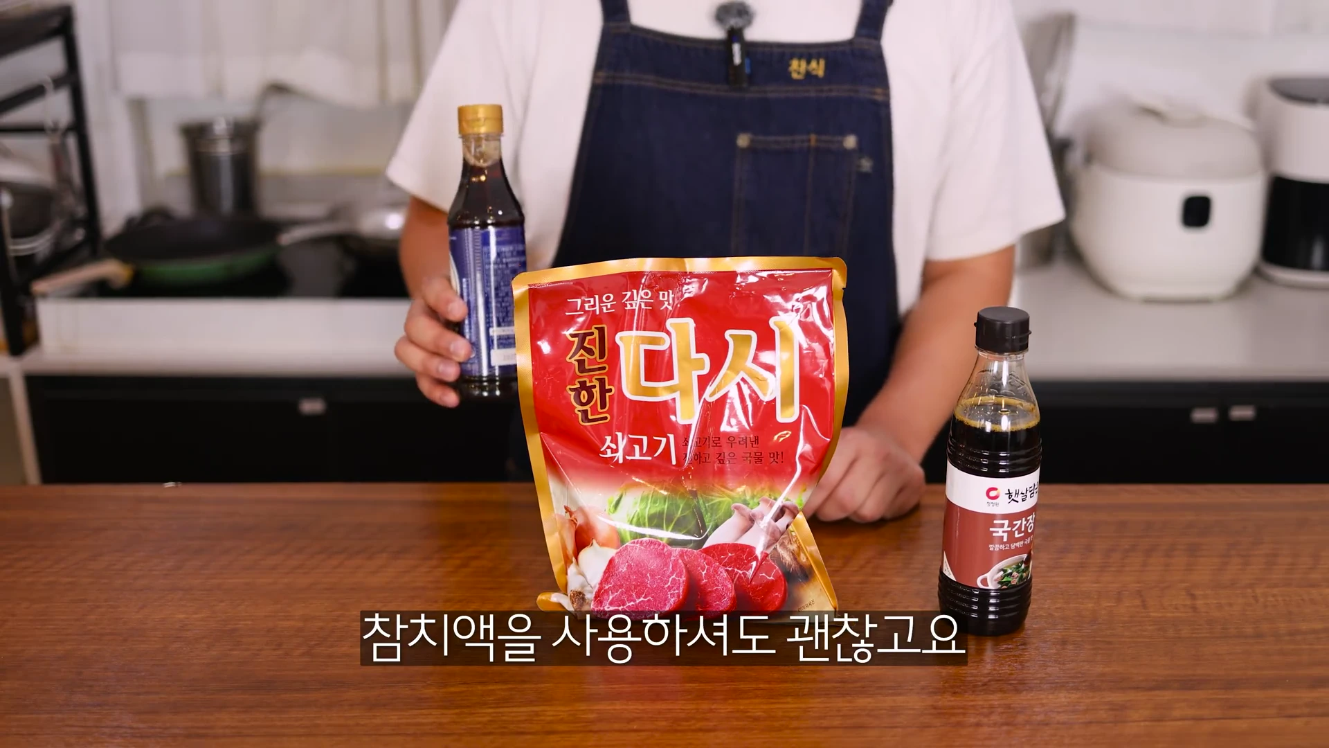 앞치마를 입은 요리사가 국간장, 다시다, 액젓 등 세 가지 양념 재료를 주방 조리대 위에 올려놓고 설명하는 모습