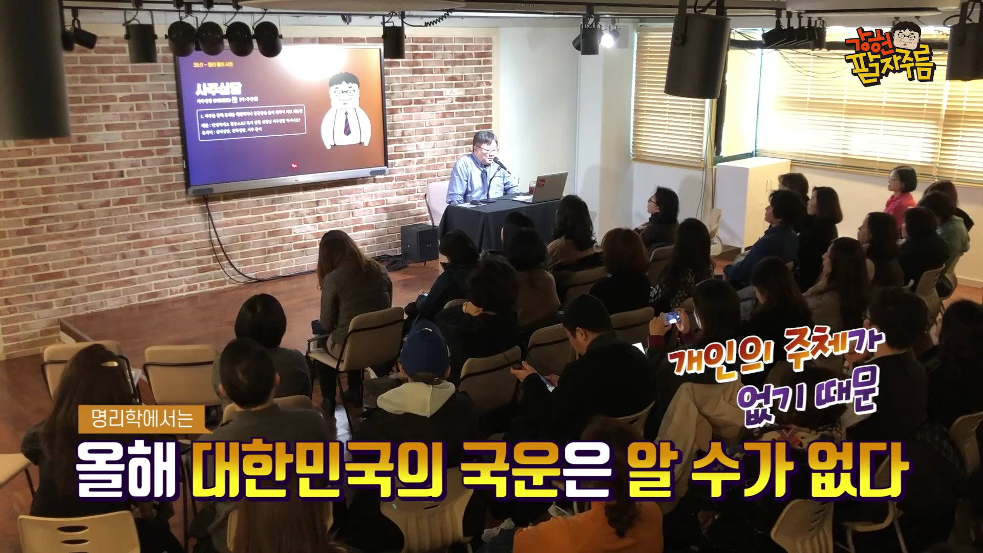 강단 앞에 앉아 마이크를 잡고 강연하는 남성과 그를 바라보는 청중들이 앉아 있는 실내 강연장 모습이며, 화면에는 '사주상담'이라는 제목의 발표 자료가 띄워져 있다.