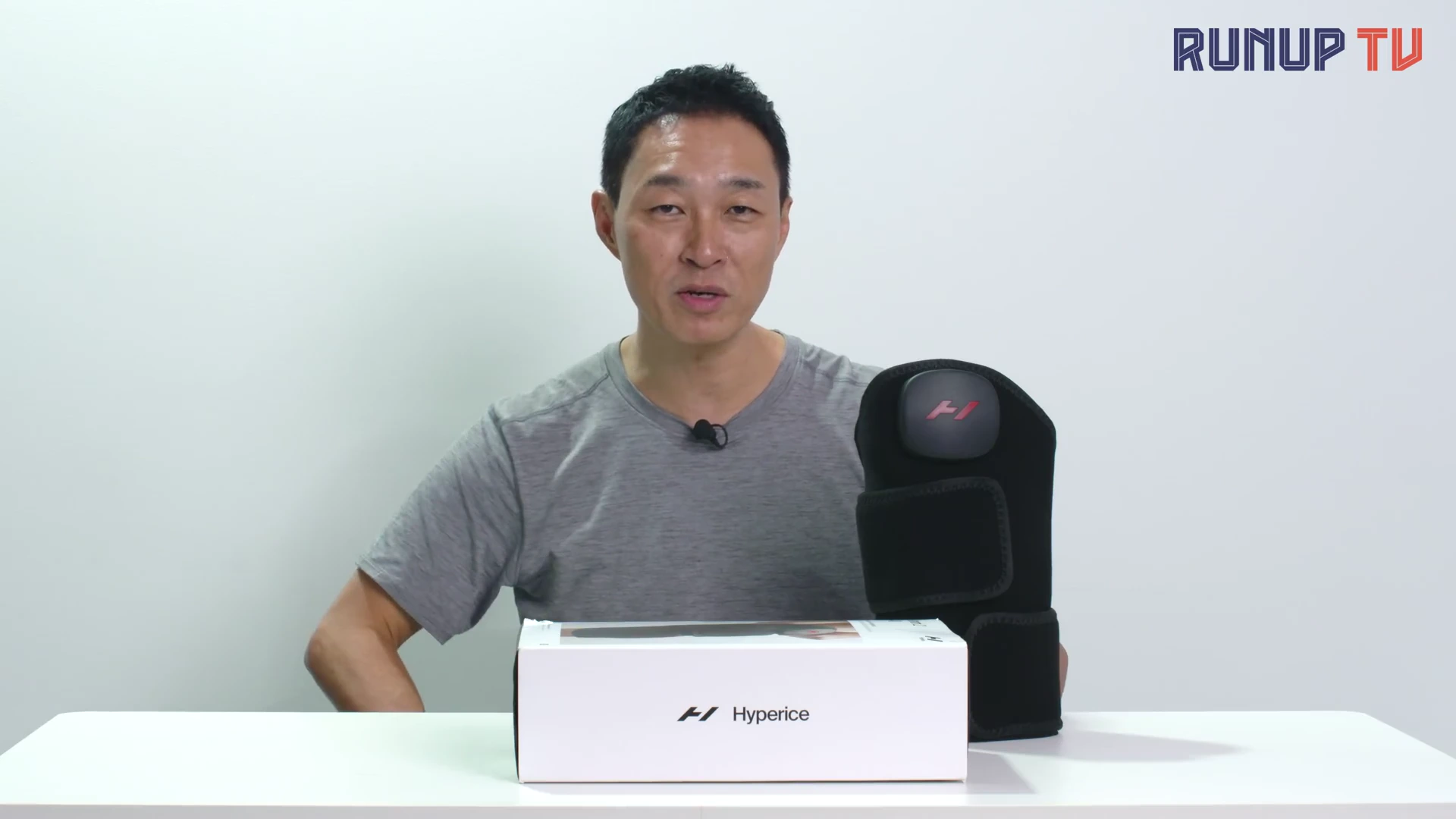 흰색 배경의 스튜디오에서 회색 티셔츠를 입은 남성이 테이블 위에 놓인 마사지기 제품과 상자를 앞에 두고 이야기를 나누고 있다.
