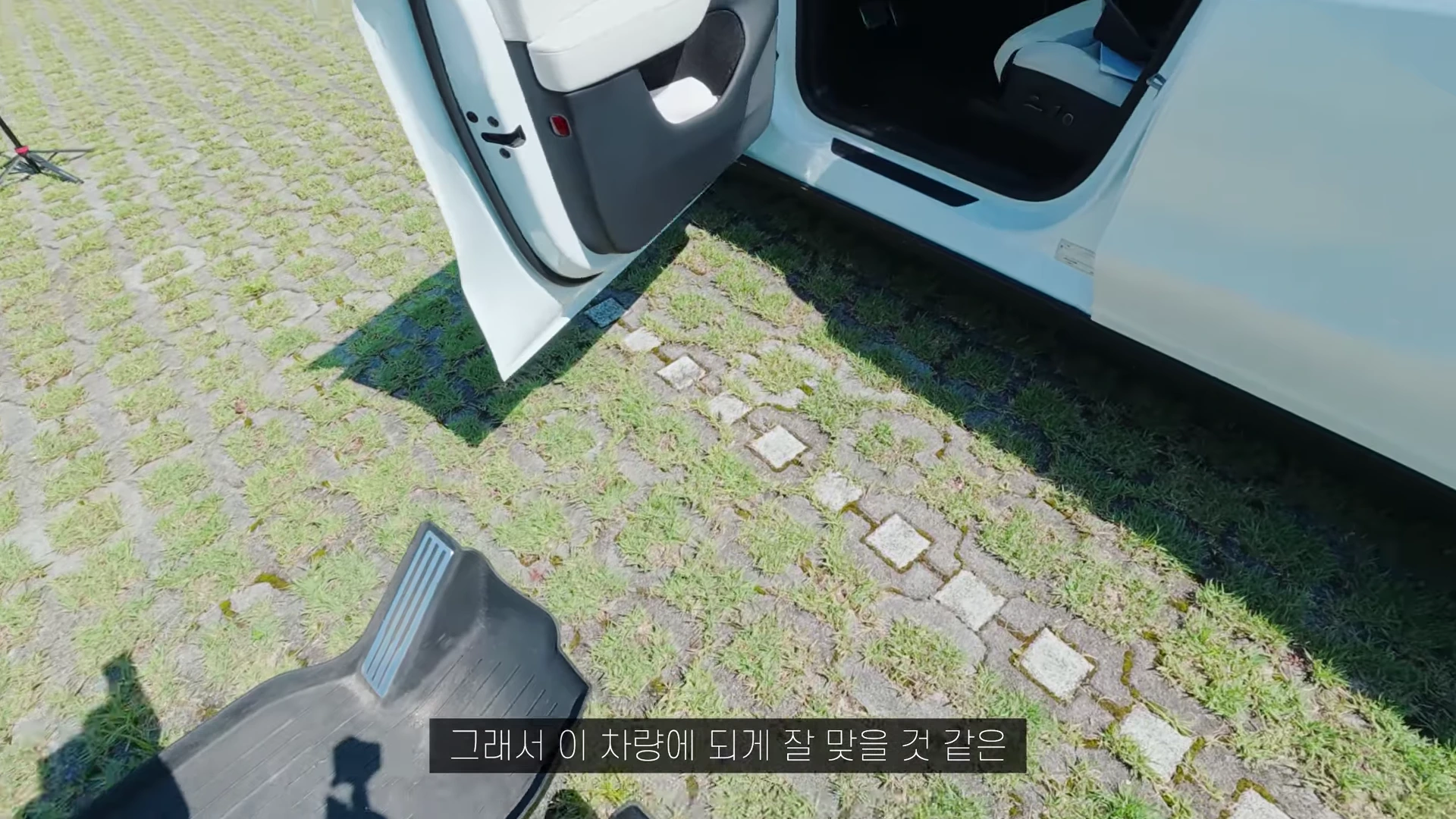 야외 주차장에 세워진 흰색 테슬라 차량의 열린 운전석 문 아래로 검은색 TPE 카매트가 놓여 있는 모습