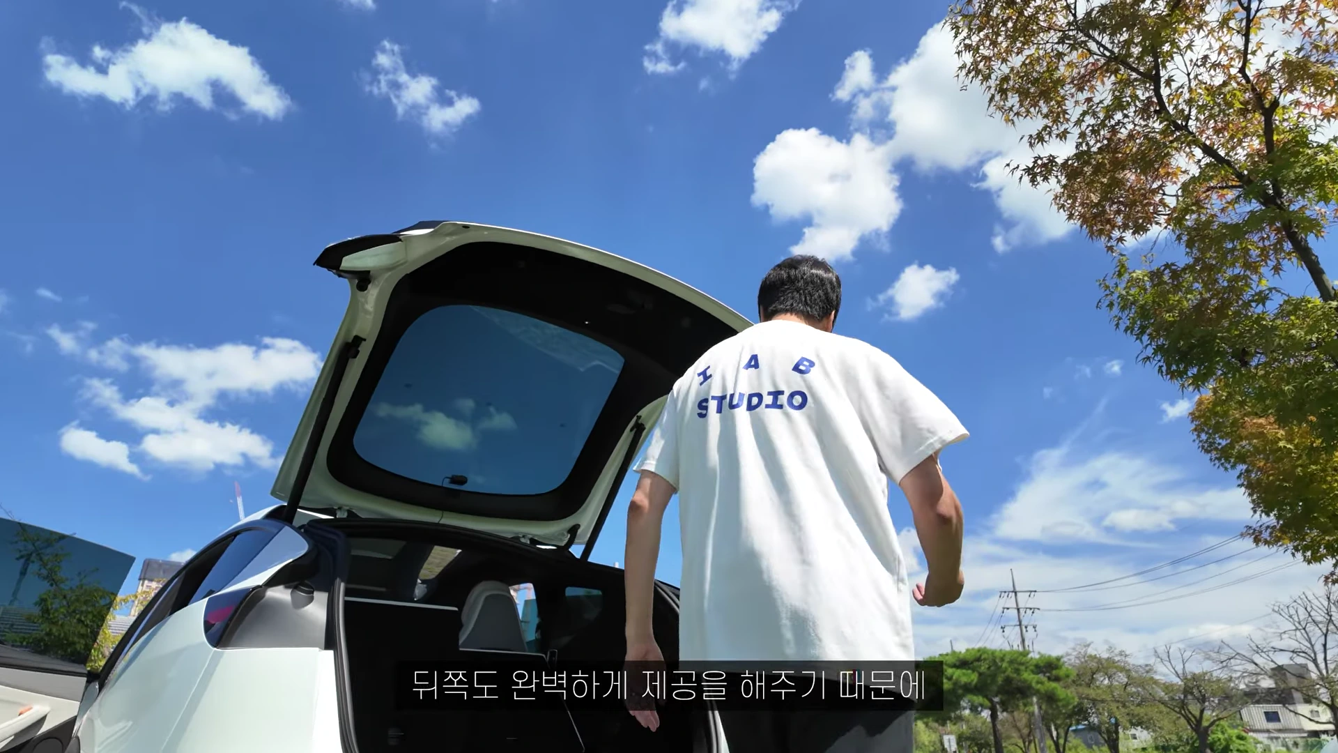 흰색 티셔츠를 입은 남성이 트렁크가 열린 흰색 자동차 뒤에서 작업을 하려고 다가가고 있으며, 배경에는 맑은 하늘과 구름이 보입니다.