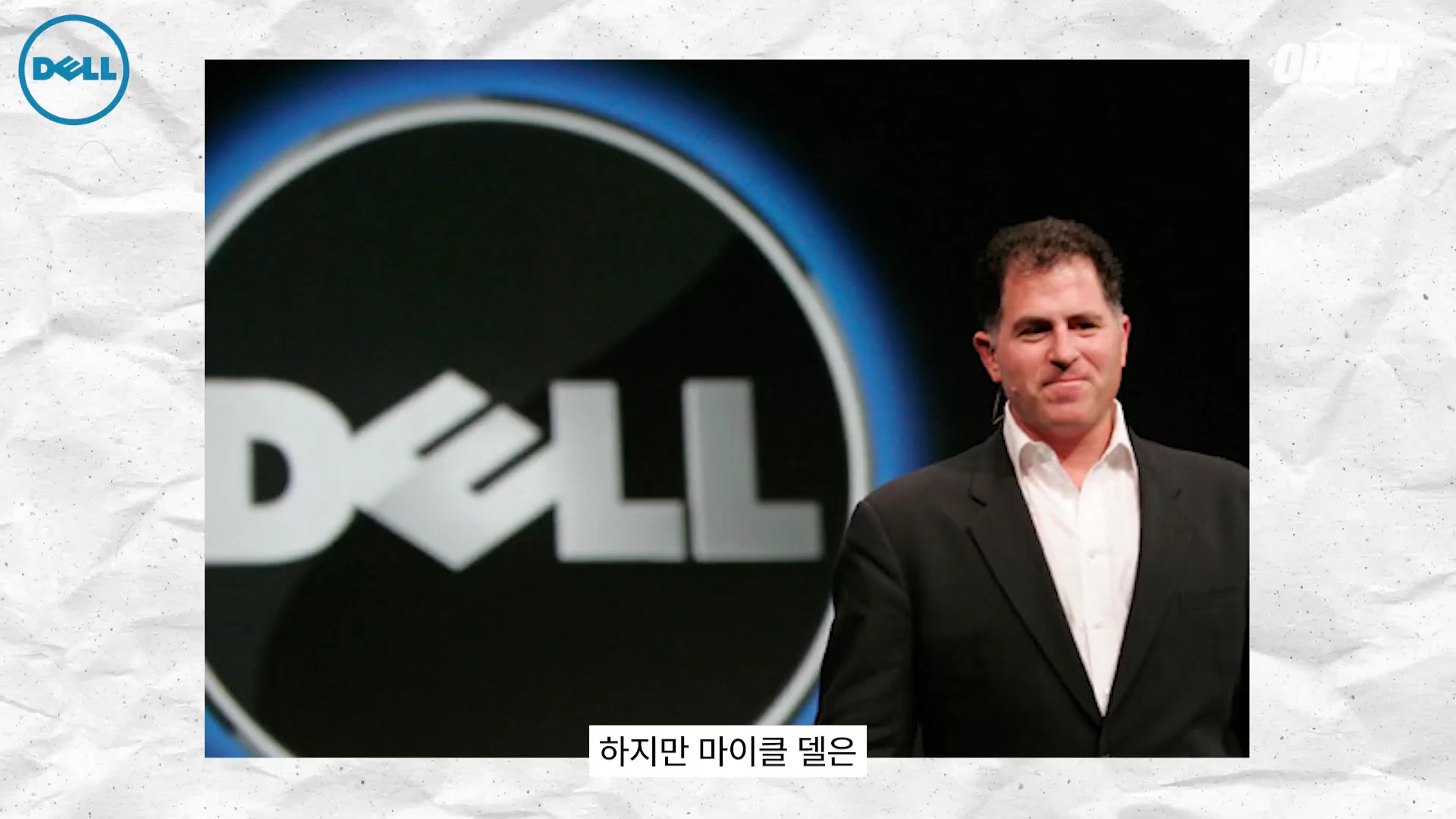 흰색 셔츠에 검은 정장을 입은 마이클 델이 대형 델 로고가 그려진 배경 앞에서 서 있는 모습