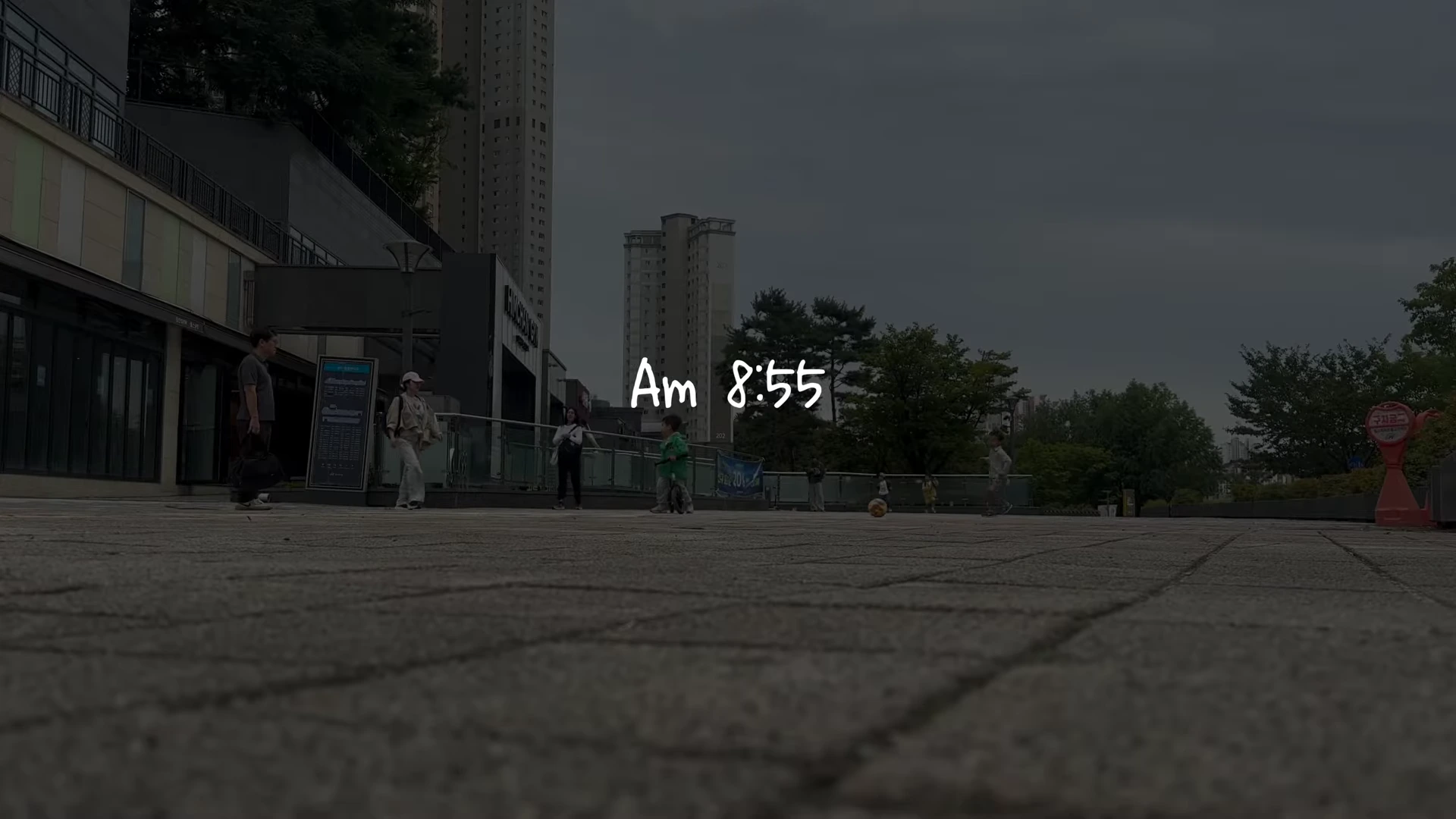 오전 8시 55분이라는 시간 텍스트가 적힌 야외 광장 풍경으로 아이들이 뛰어놀고 주변에는 건물과 나무가 보인다.