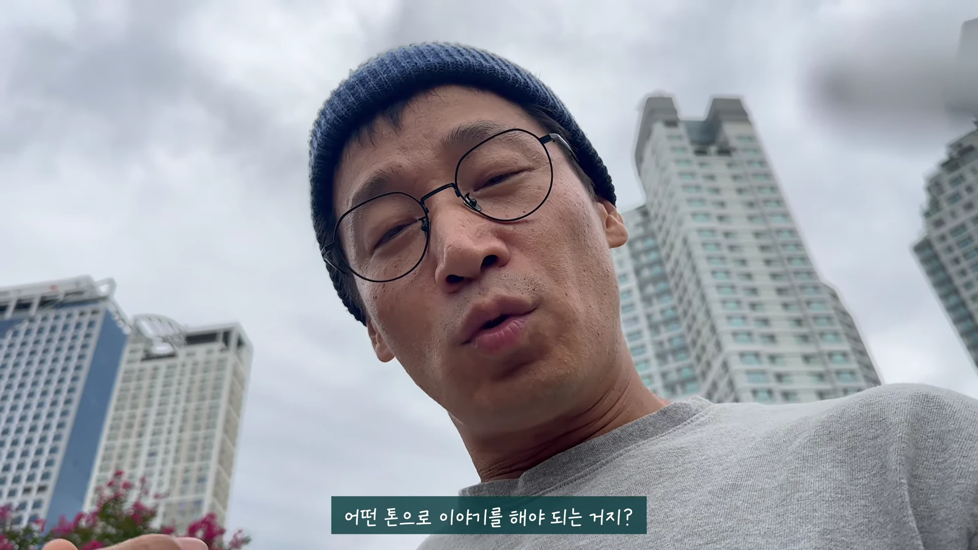 비니를 쓰고 안경을 쓴 남성이 야외에서 카메라를 내려다보며 무언가 말하고 있다.