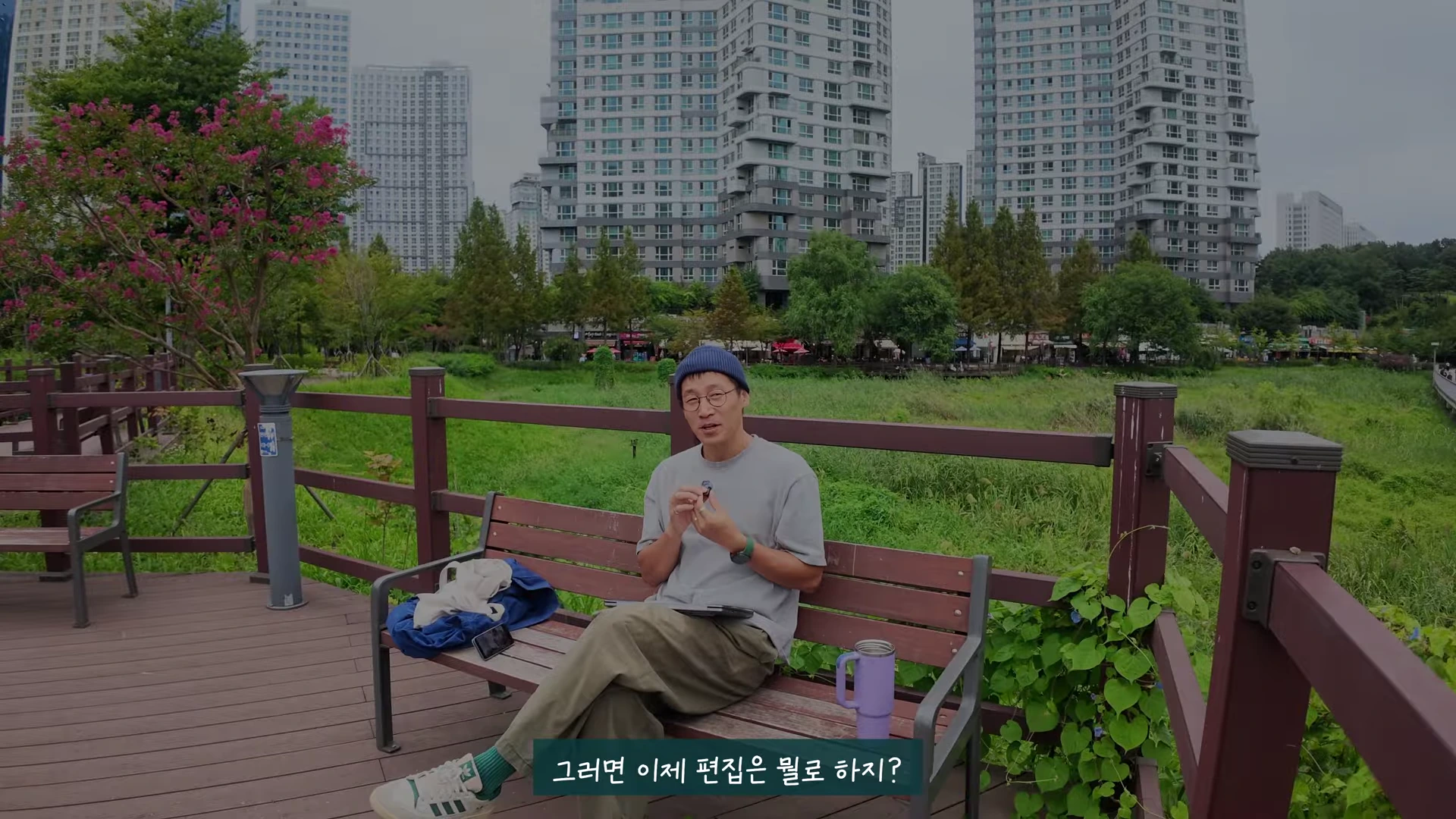안경을 쓰고 비니를 쓴 남성이 호수공원의 나무 벤치에 앉아 무언가를 설명하며 이야기를 하고 있다.