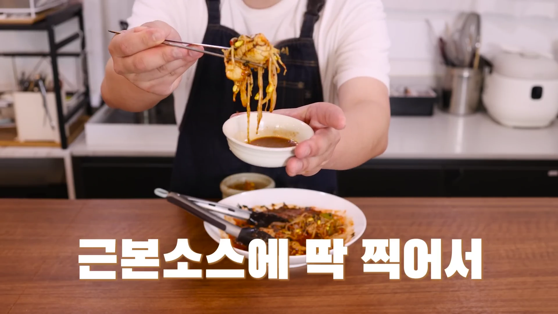 앞치마를 입은 요리사가 젓가락으로 콩나물과 명란이 섞인 요리를 집어 간장 소스에 찍으려는 모습이 담긴 클로즈업 영상 캡처 화면이며, 하단에는 '근본소스에 딱 찍어서'라는 자막이 있습니다.