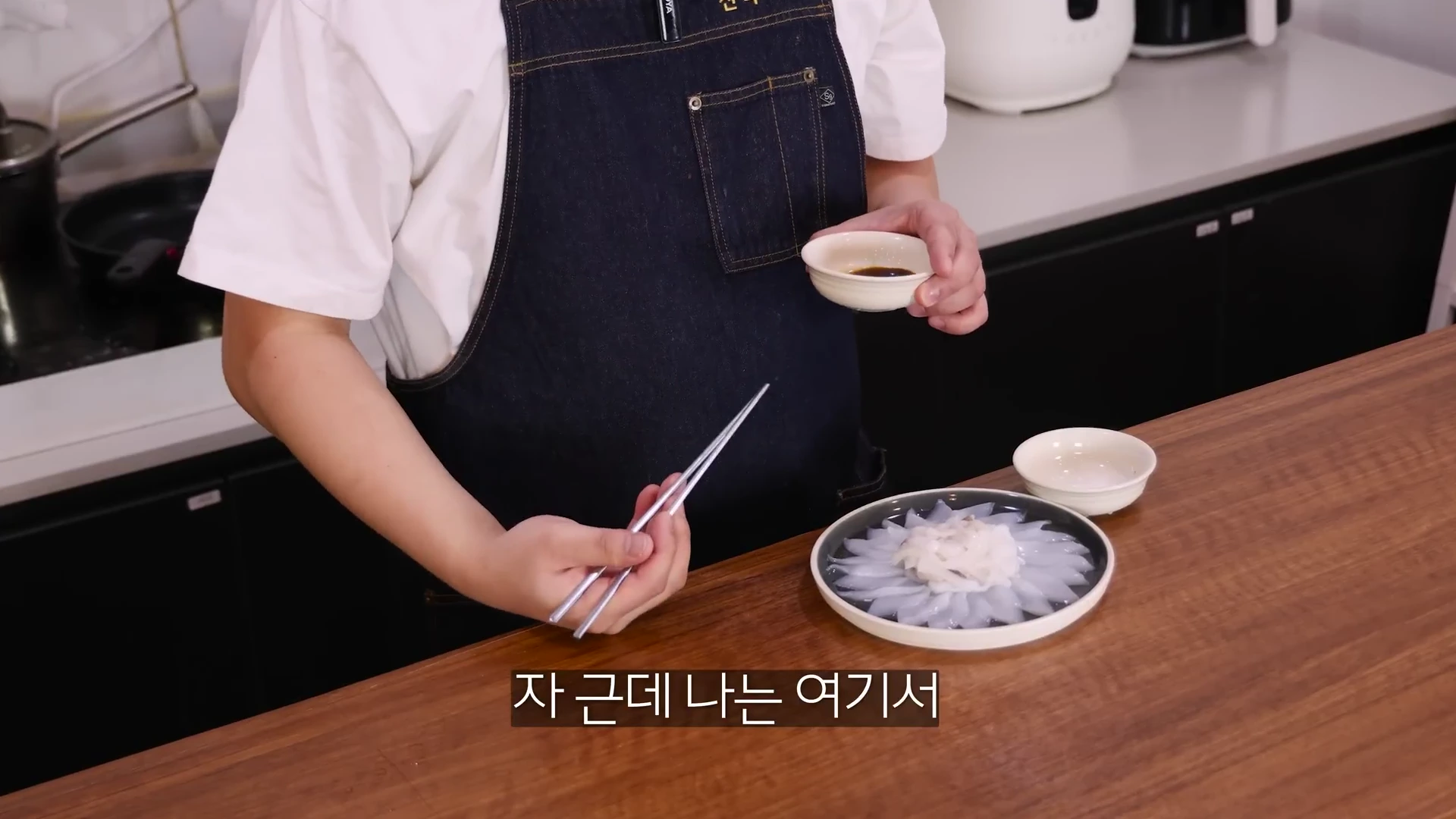 앞치마를 두른 한 사람이 젓가락과 작은 소스 종지를 들고 꽃 모양으로 정갈하게 놓인 오징어 회 접시 옆에 서 있는 모습