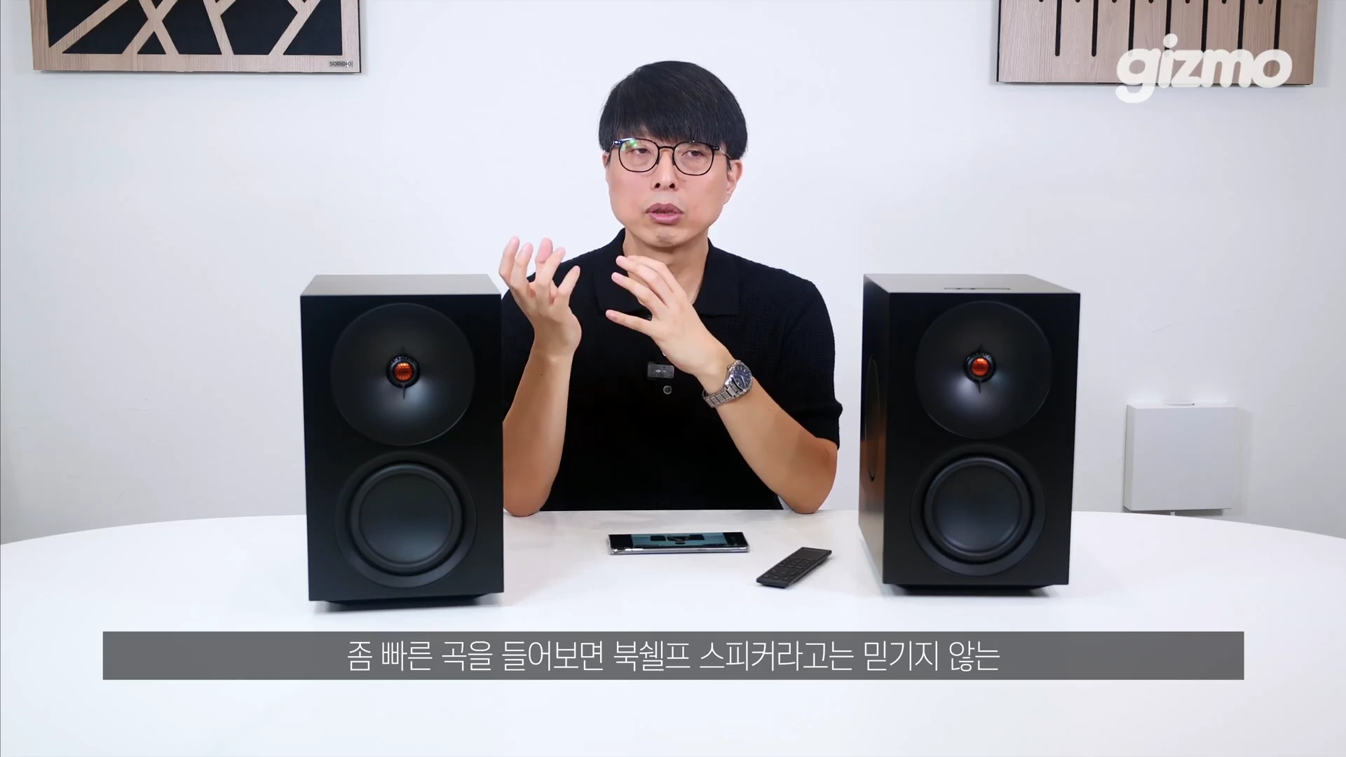 안경을 쓴 남성이 흰색 테이블 위에 놓인 검은색 스피커 두 대 사이에 앉아 손을 들어 설명하고 있는 모습.