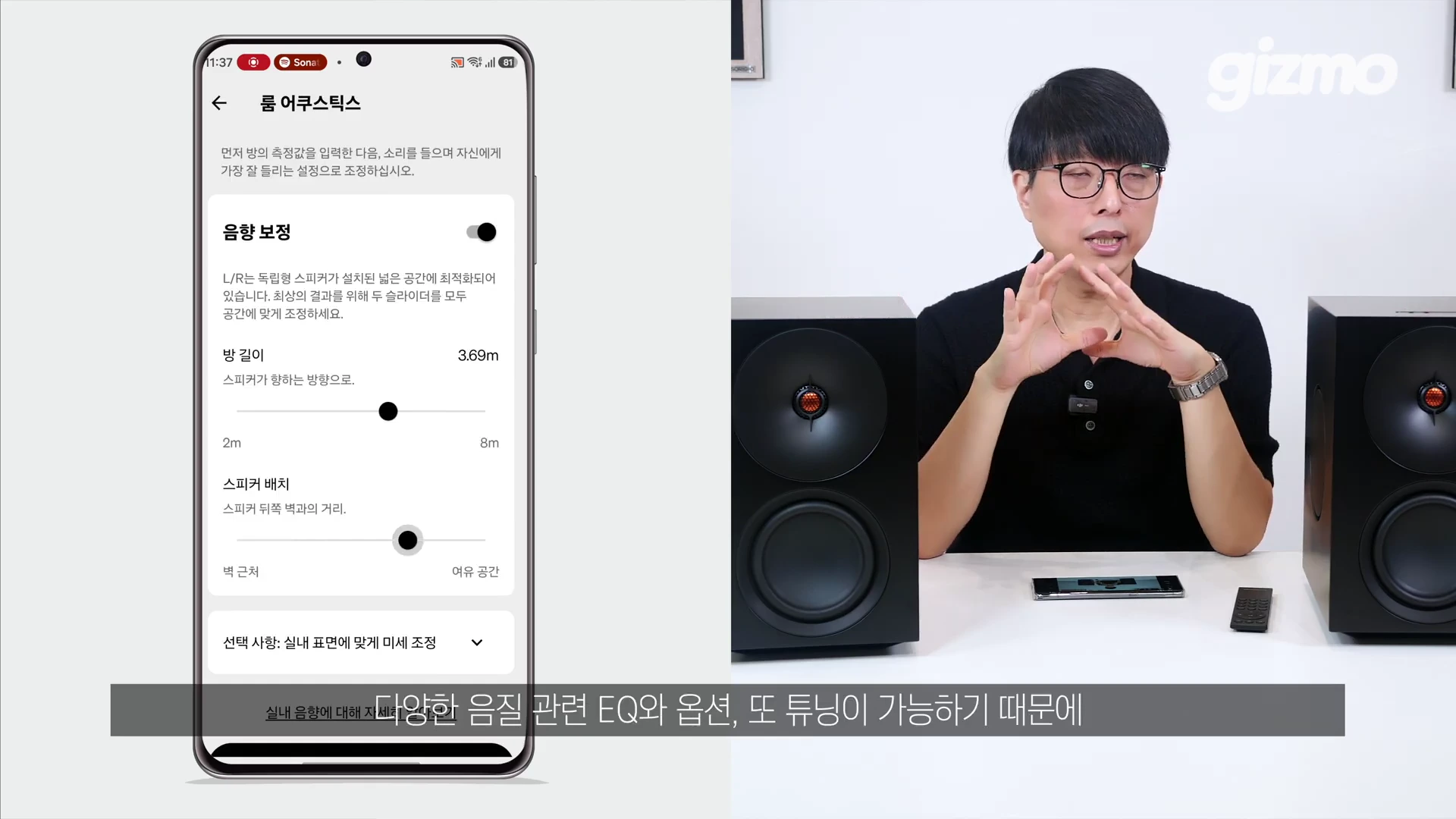 안경을 쓴 남성이 스피커 두 대 사이에 앉아 손짓하며 설명하고 있고, 왼쪽에는 모바일 앱 화면에 룸 어쿠스틱 설정 페이지가 표시되어 있습니다.