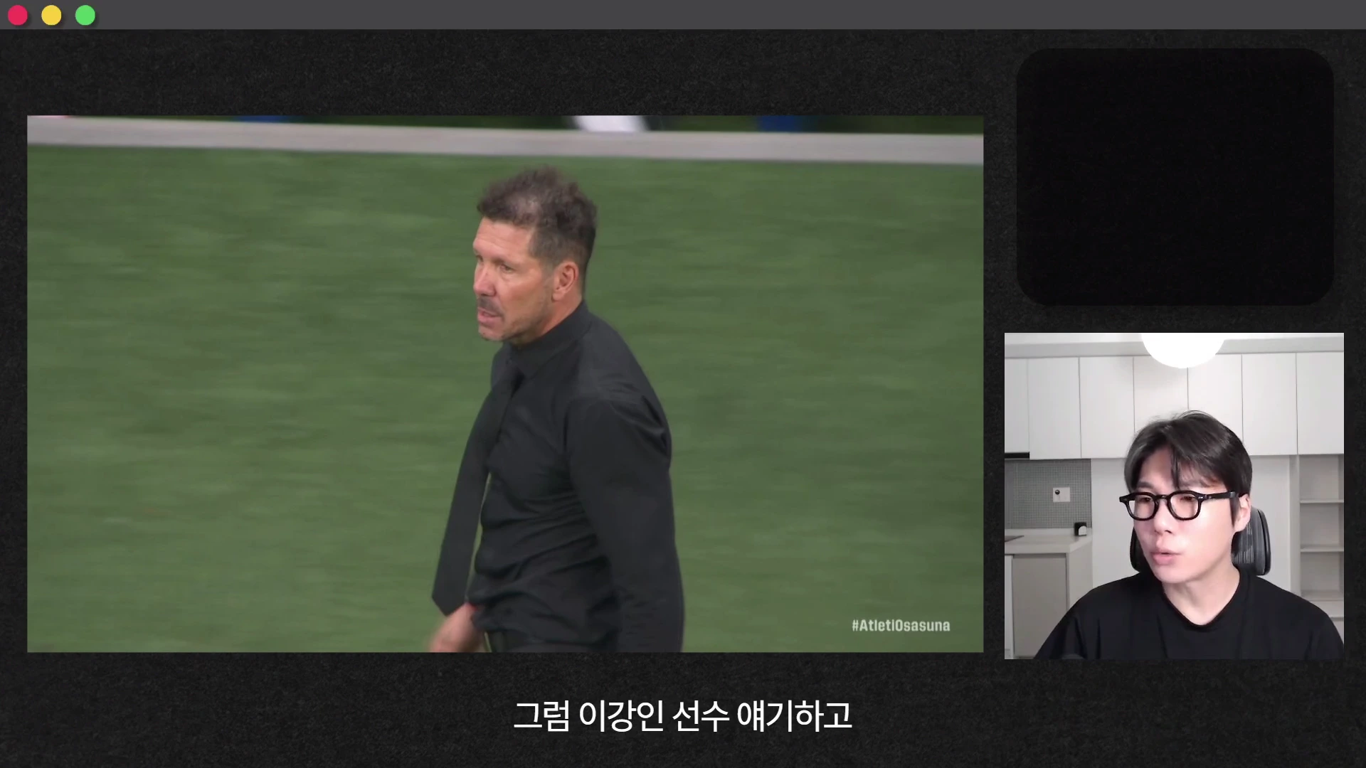 축구 경기장에서 검은 옷을 입고 서 있는 감독과 그 옆에 안경을 쓴 채 이야기하는 남성이 화면에 함께 나타나 있는 영상 프레임입니다.