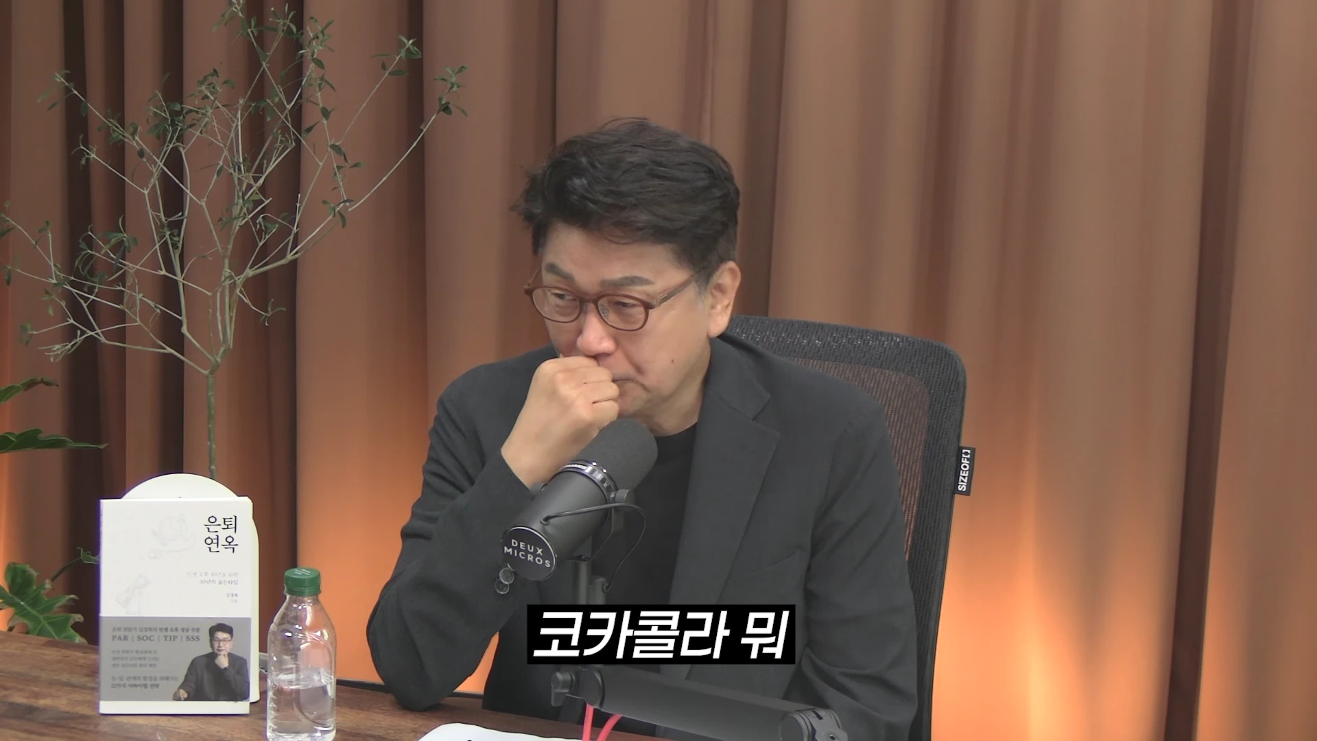 안경을 쓴 중년 남성이 테이블 앞에 앉아 마이크에 대고 이야기하고 있으며, 테이블 위에는 책 한 권과 물병이 놓여 있고 화면 하단에는 코카콜라 뭐라는 자막이 떠 있다.