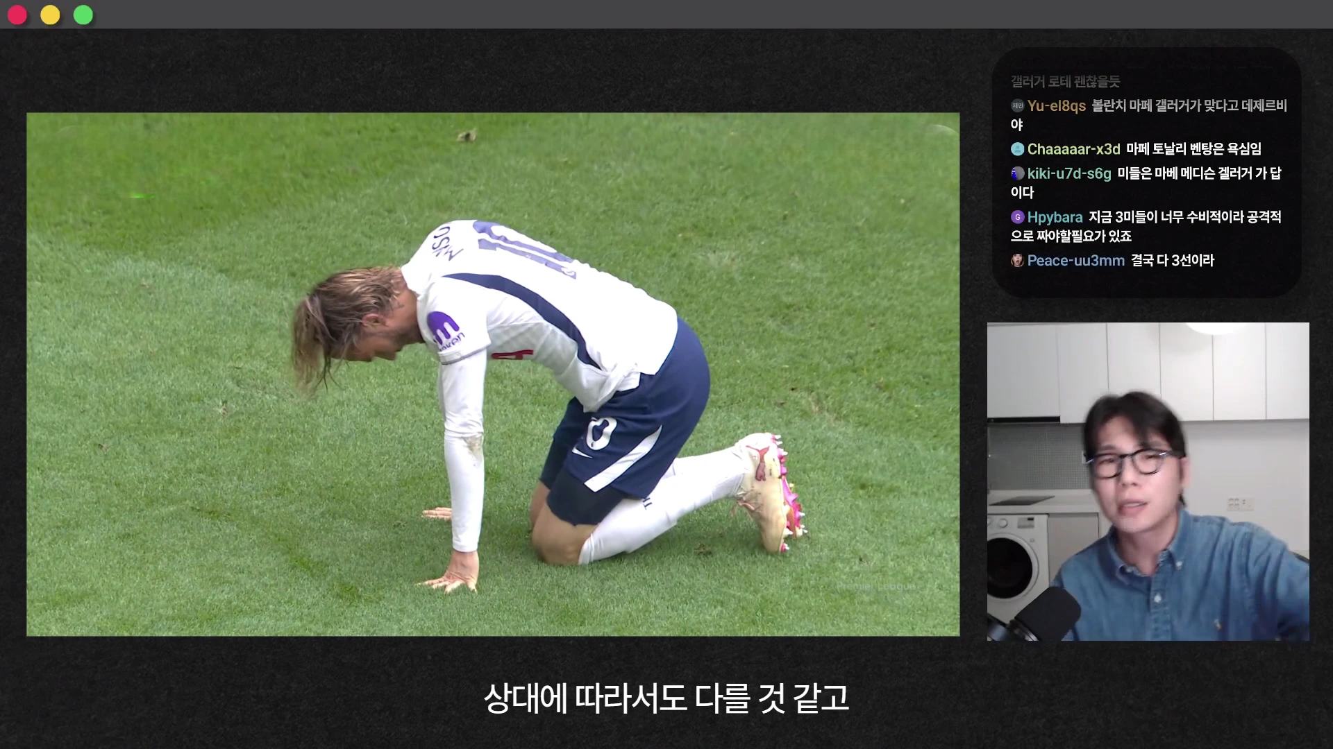 축구 경기장에서 한 선수가 잔디 위에 무릎을 꿇고 고개를 숙이고 있으며, 화면 오른쪽에는 안경을 쓴 남성이 마이크 앞에서 이야기하는 모습과 실시간 채팅창이 포함된 영상 화면.