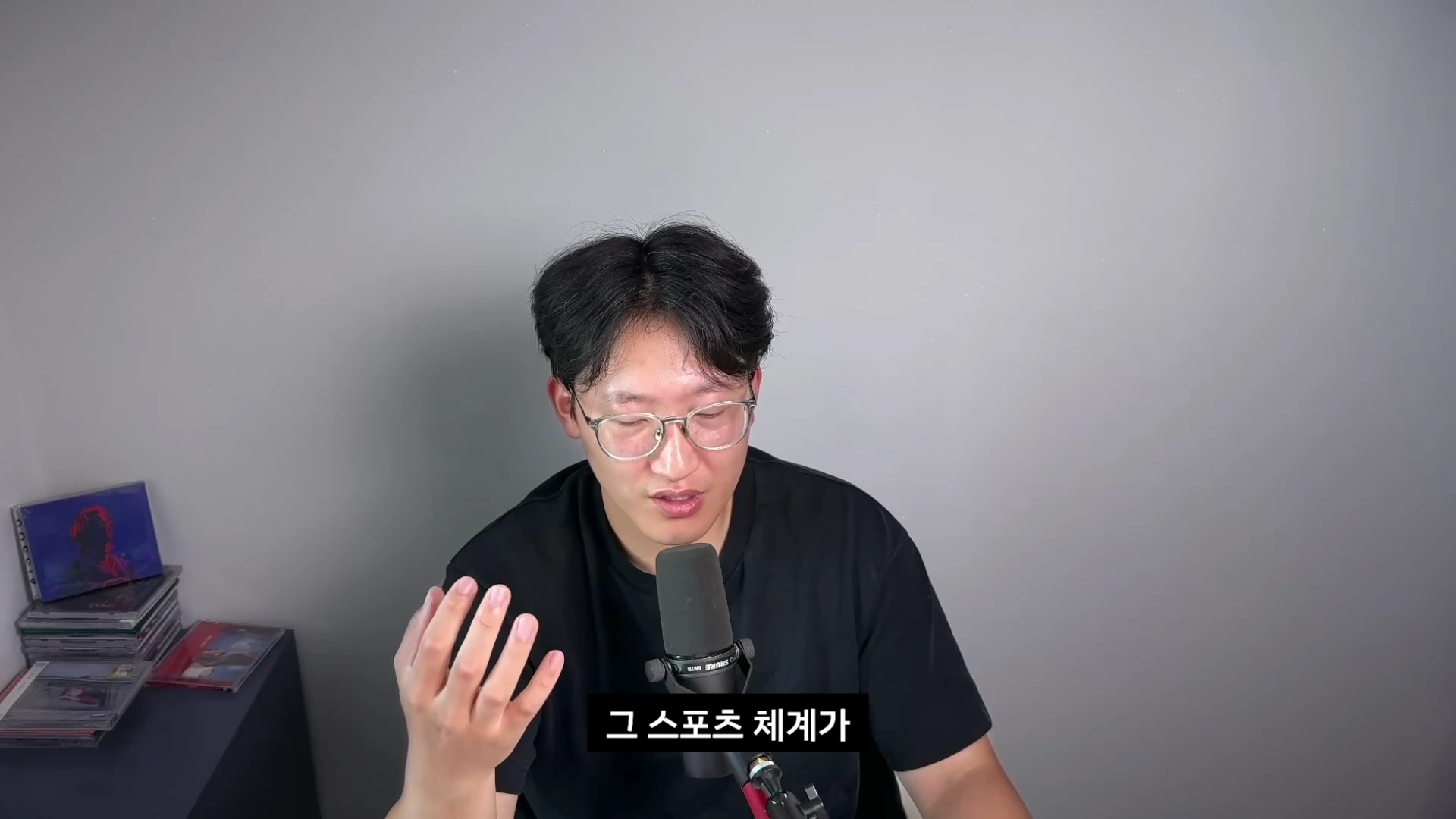 안경을 쓴 남성이 마이크 앞에서 이야기하고 있으며 하단에 '그 스포츠 체계가'라는 자막이 표시된 화면.
