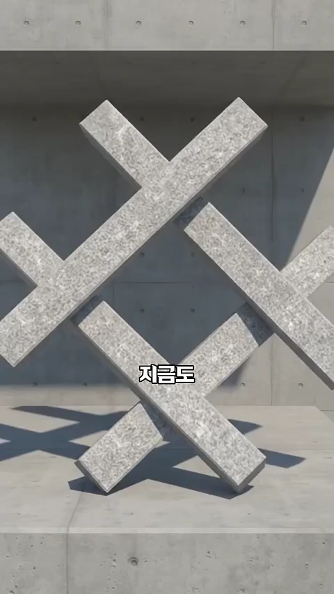 콘크리트 벽 앞에 X자 형태로 교차된 석재 구조물이 설치되어 있고 화면 중앙에는 '지금도'라는 자막이 배치되어 있습니다.