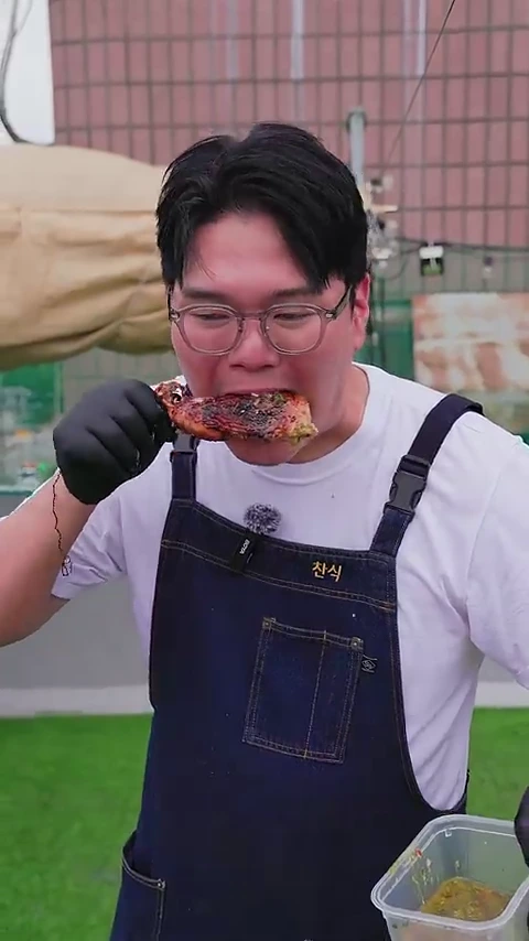 안경을 쓴 남성이 검은색 위생 장갑을 끼고 구운 닭고기 한 점을 입으로 베어 물고 있는 모습