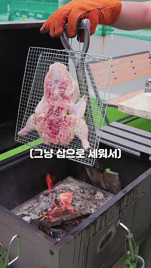 주황색 작업용 장갑을 낀 손이 납작하게 펼쳐진 생닭을 고정시킨 철제 석쇠를 들고 화로 옆에 세워두고 있는 모습입니다.