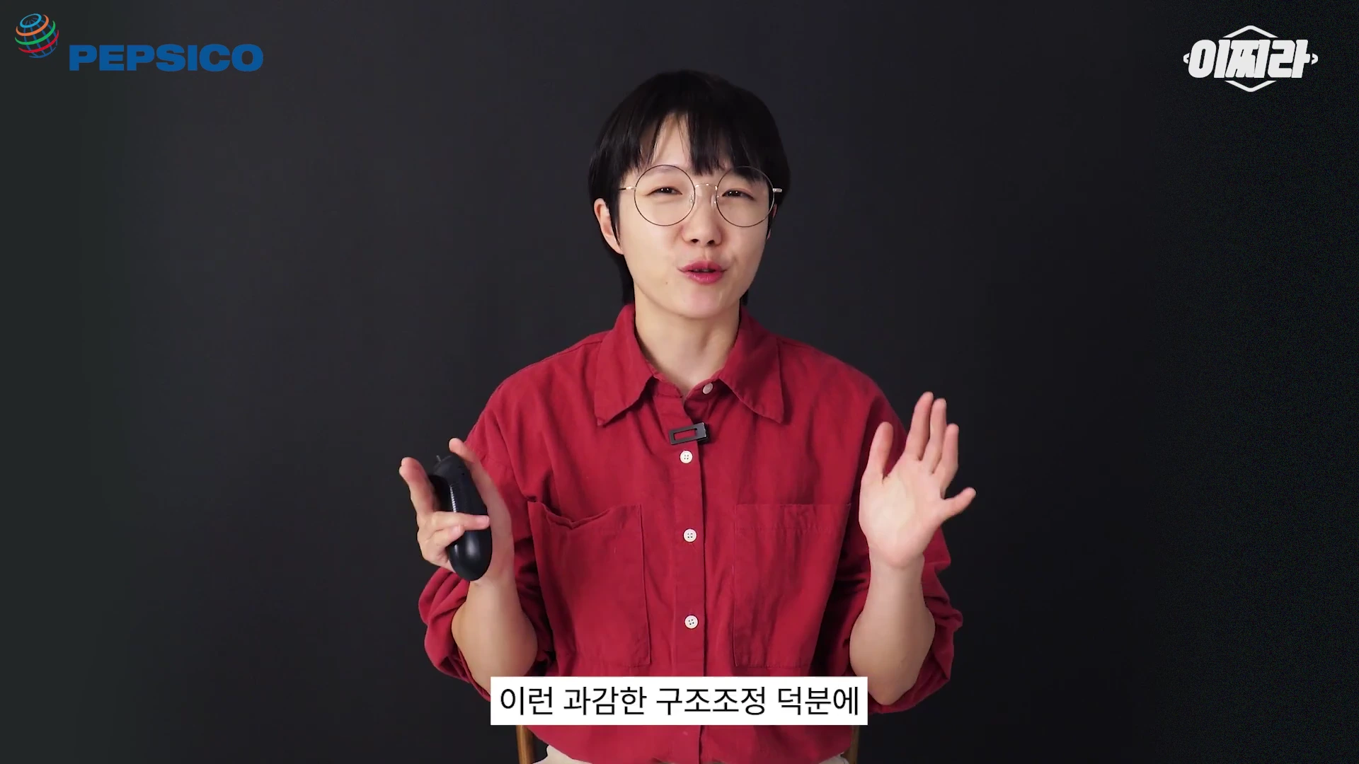 단발머리에 안경을 쓴 진행자가 붉은색 셔츠를 입고 검은색 배경 앞에서 손동작을 하며 이야기하고 있다.