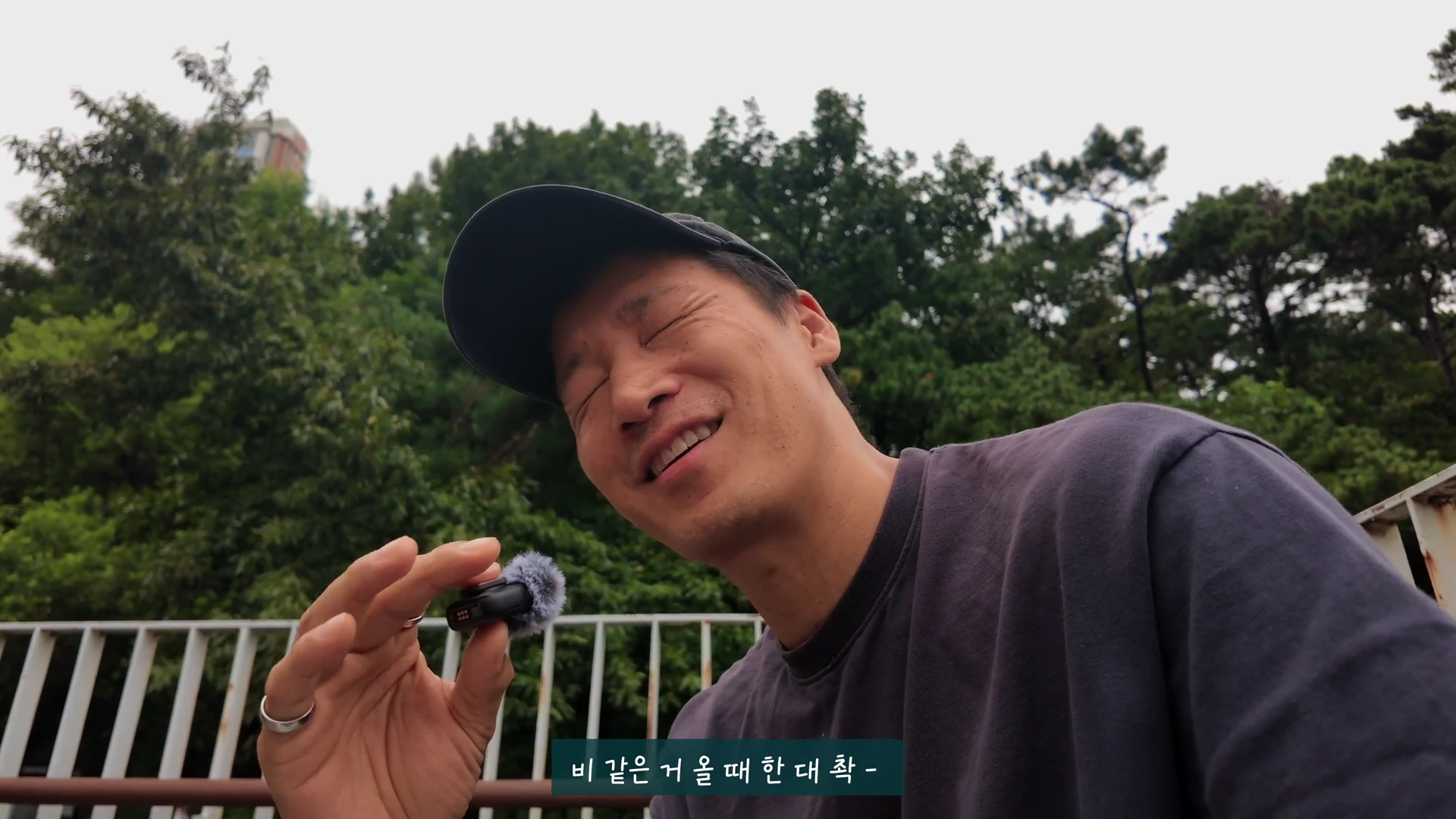 검은 모자를 쓴 남성이 야외에서 웃으며 손에 작은 무선 마이크를 들고 이야기하고 있다.