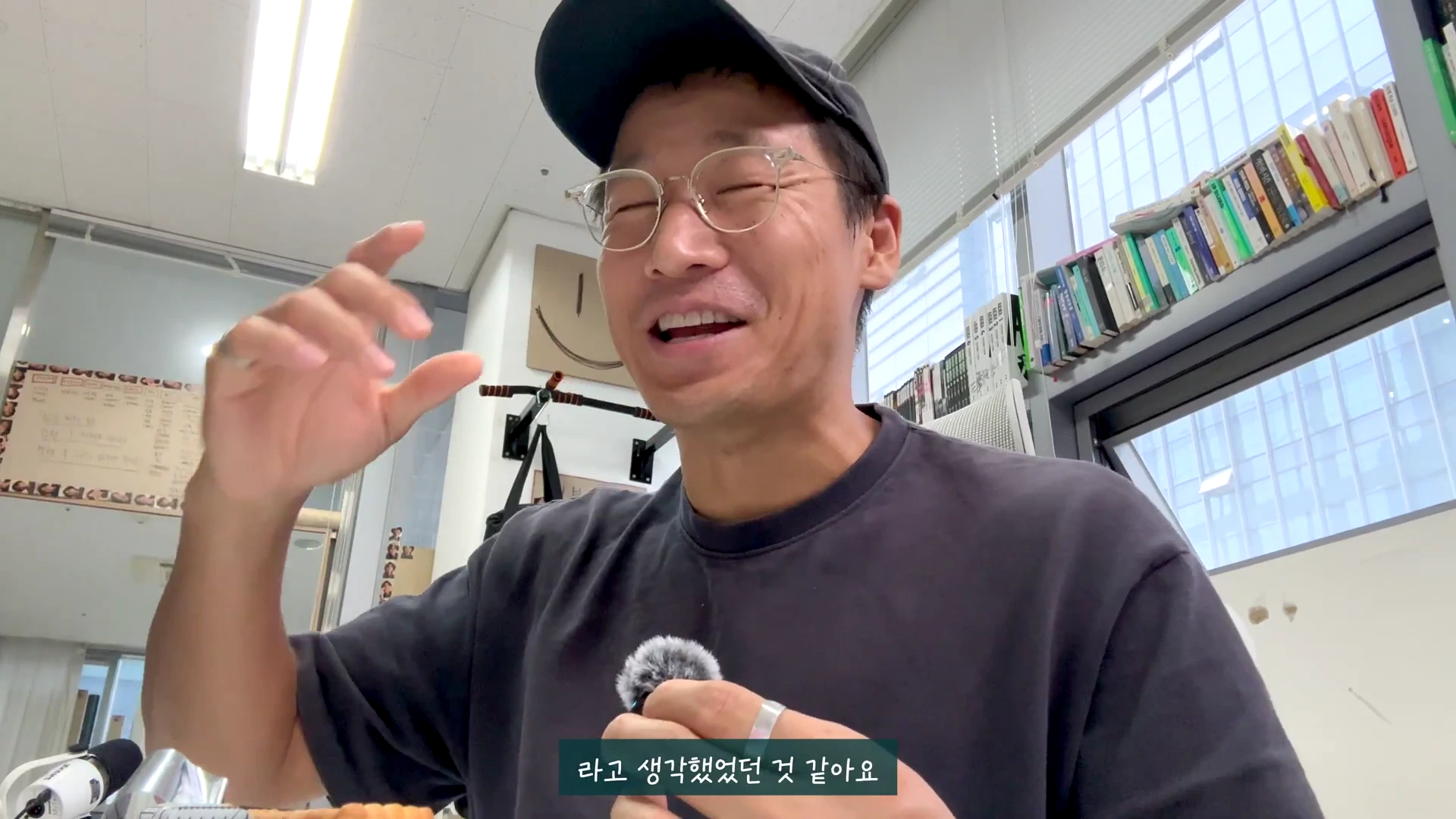 실내에서 검은색 티셔츠와 야구 모자를 쓴 남성이 환하게 웃으며 손짓으로 이야기를 나누고 있다. 뒤편에는 책장이 있고, 하단에는 자막이 떠 있는 영상 화면이다.