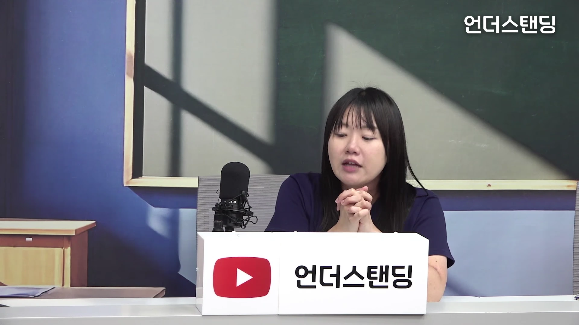 어두운 남색 상의를 입은 여성이 책상 앞에 앉아 손을 맞잡고 이야기를 나누고 있으며, 앞에는 로고가 새겨진 상자가 놓여 있고 배경에는 칠판이 있는 스튜디오 공간