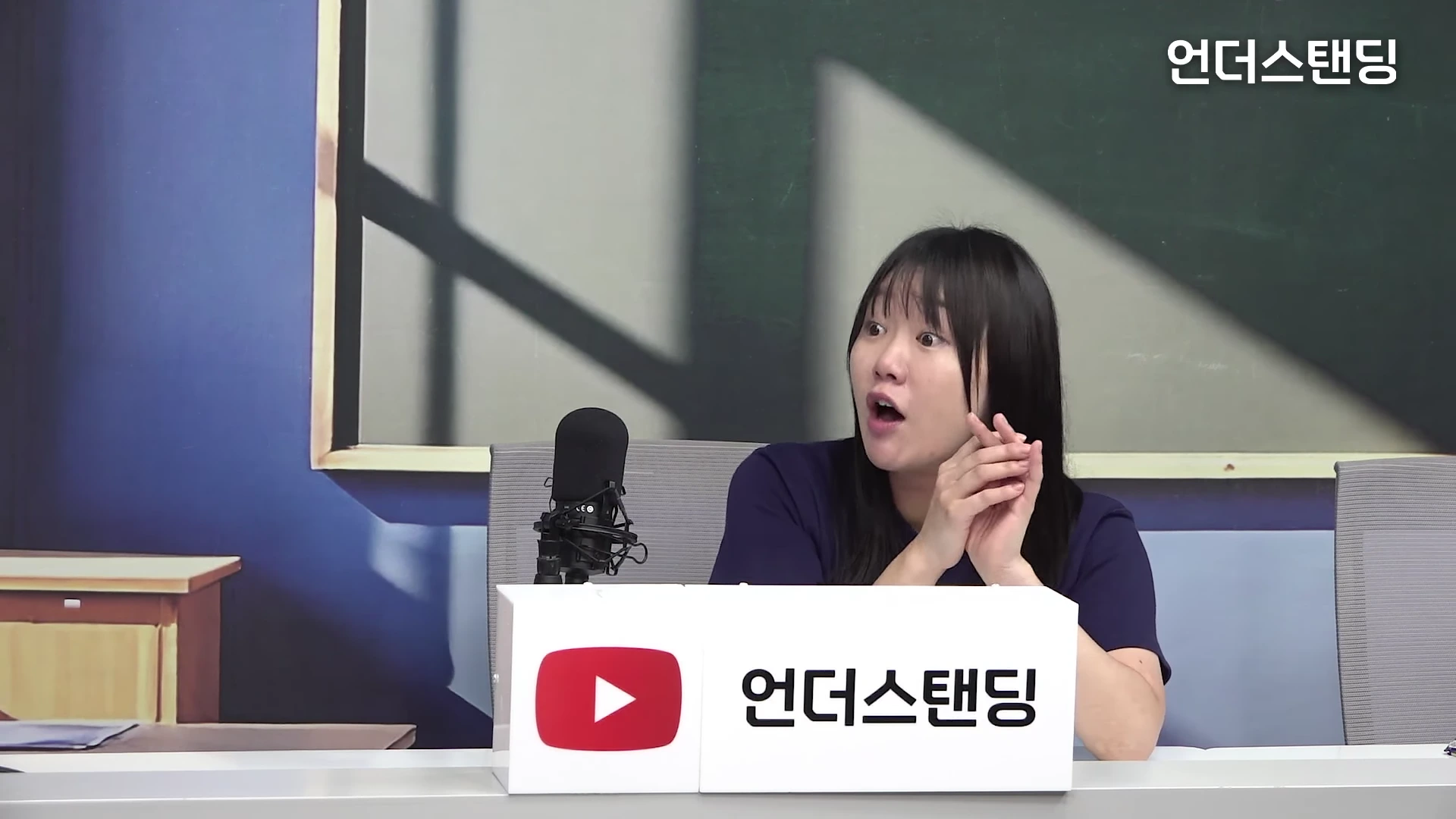 스튜디오 의자에 앉아 있는 여성이 마이크 앞에서 손을 모으고 진지하게 이야기를 나누고 있으며, 앞쪽에는 유튜브 로고가 그려진 상자가 놓여 있습니다.