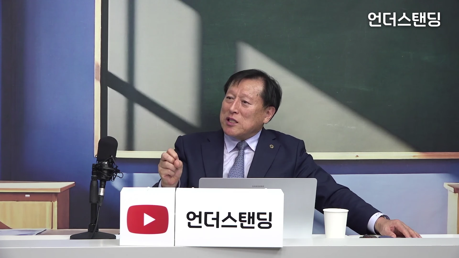 정장을 입은 중년 남성이 스튜디오 테이블에 앉아 마이크 앞에 대고 손을 들어 이야기를 나누고 있는 모습