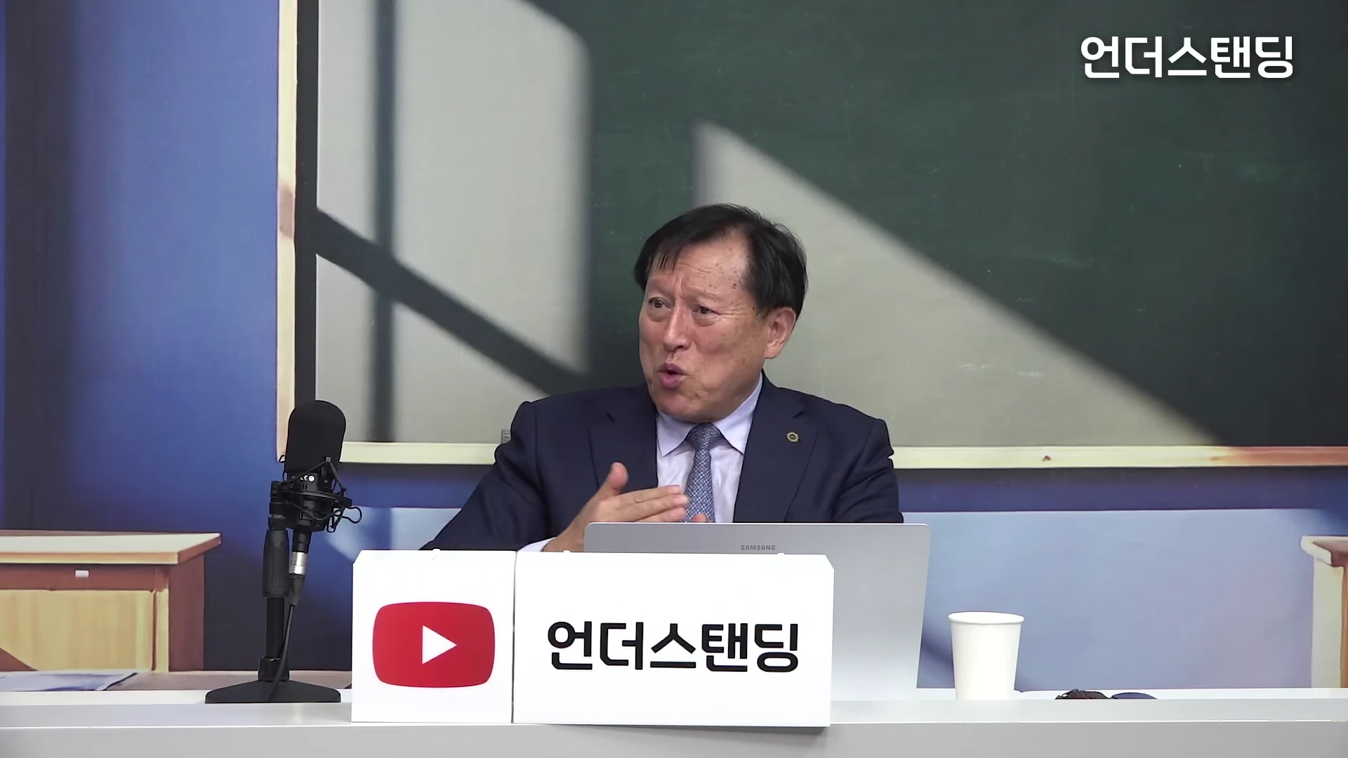 정장을 입은 중년 남성이 스튜디오 책상 앞에 앉아 마이크를 향해 손동작을 하며 이야기하는 모습