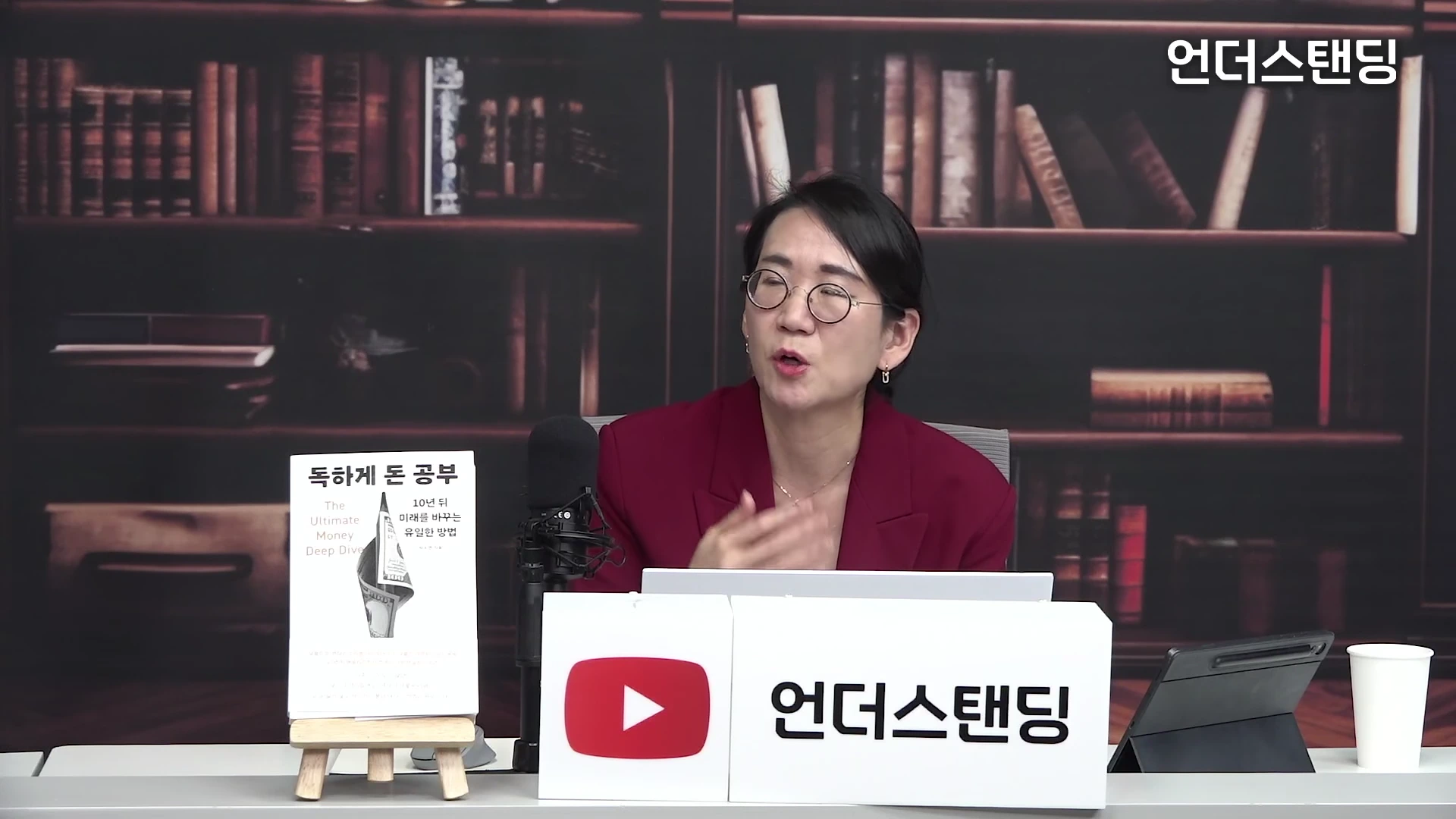 서재 배경의 스튜디오에서 안경을 쓴 여성이 앉아 이야기를 나누고 있으며, 앞쪽에는 '독하게 돈 공부'라는 제목이 적힌 책자와 로고가 놓여 있습니다.