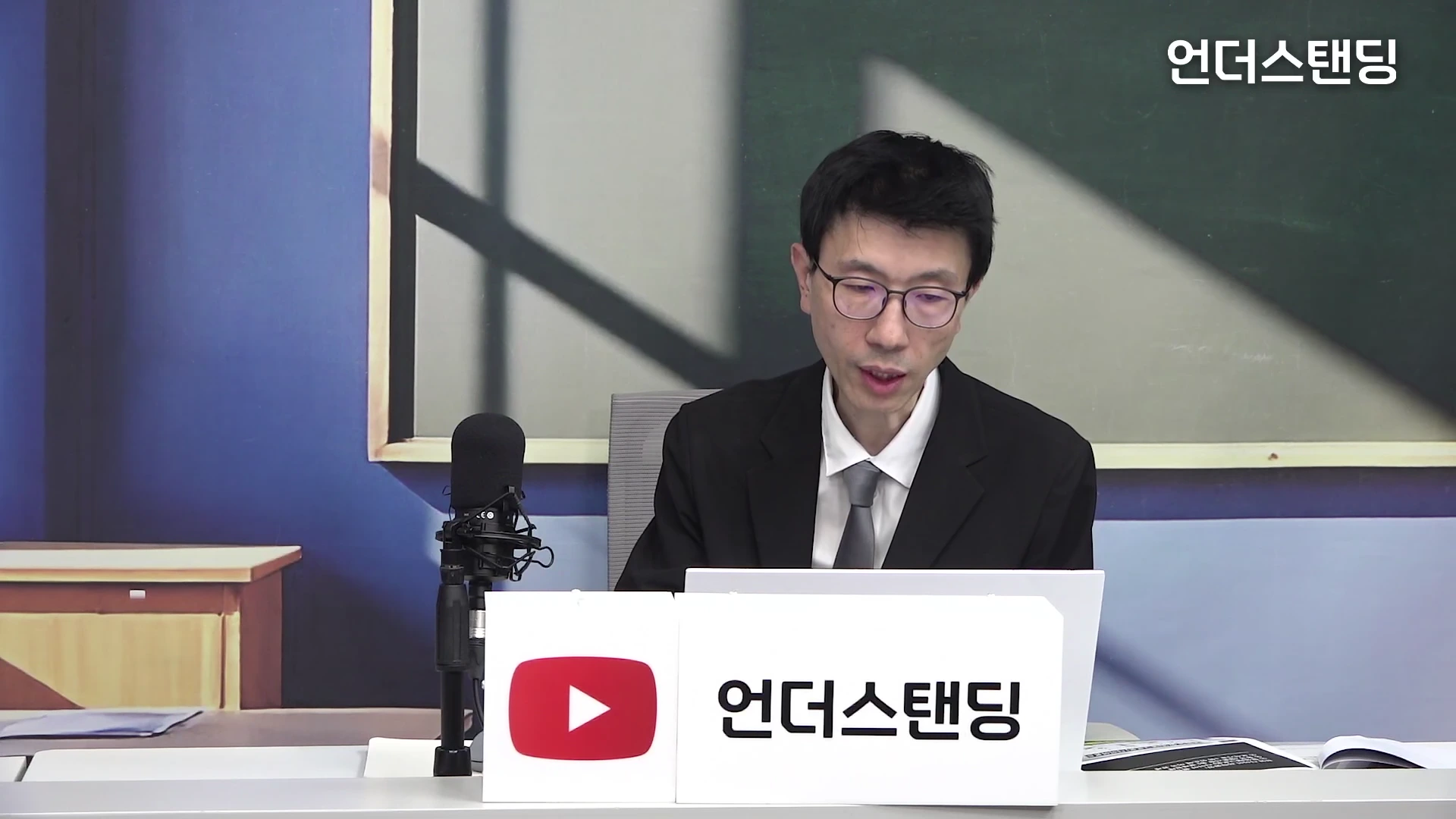 안경을 쓴 남성이 스튜디오 책상에 앉아 노트북을 보며 이야기를 하고 있고 앞에는 마이크와 채널 이름이 적힌 명패가 놓여 있습니다.