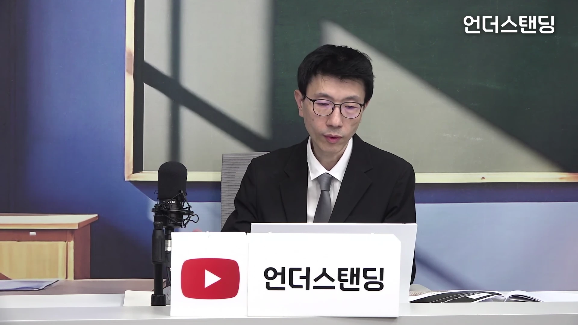 한 남성이 스튜디오 책상에 앉아 노트북을 보며 이야기하고 있고, 앞에는 마이크와 '언더스탠딩' 로고가 놓여 있습니다.