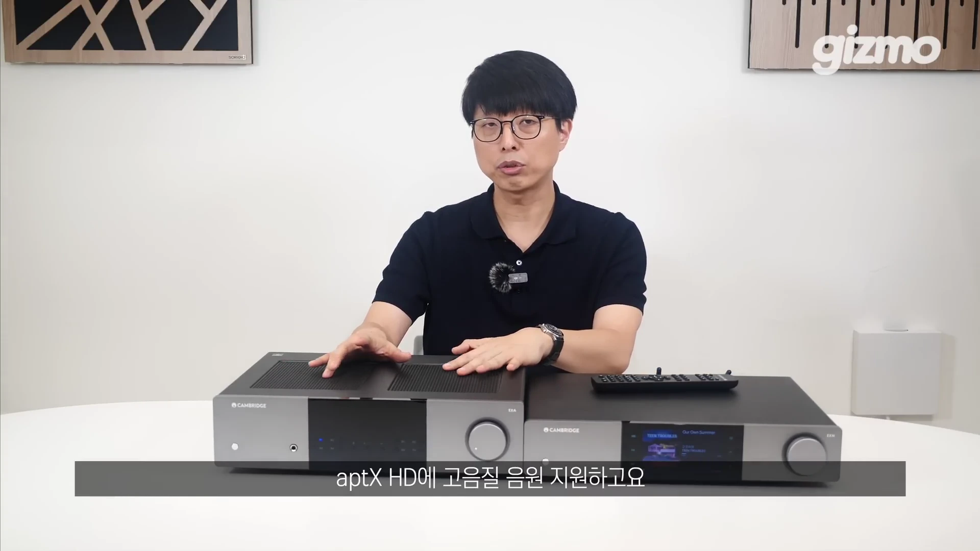 안경을 쓴 남성이 테이블 위에 놓인 캠브리지 오디오 인티앰프와 네트워크 스트리머 옆에서 손을 올린 채 설명하고 있는 스튜디오 인터뷰 장면.