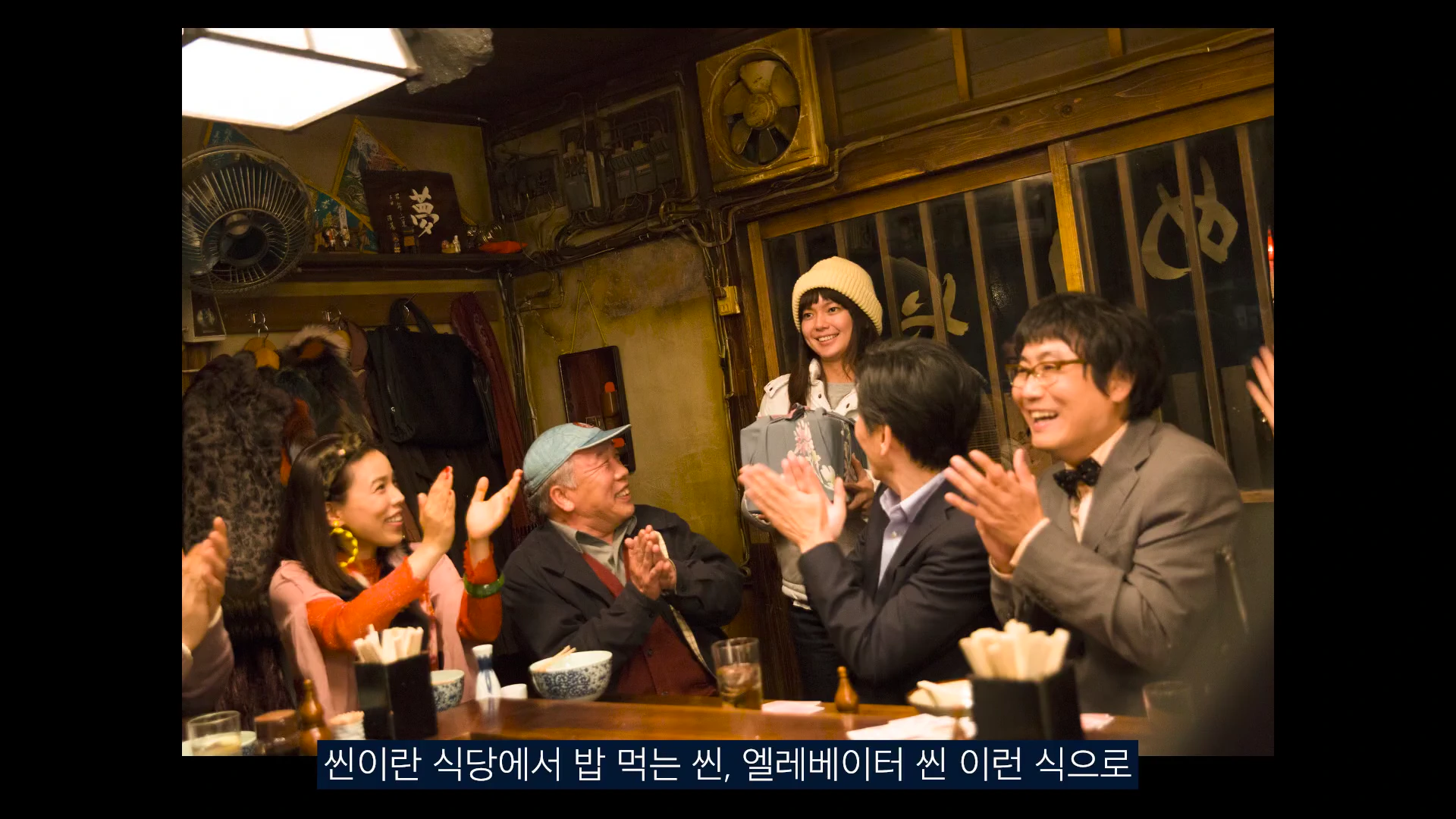 식당 안에서 여러 사람이 테이블에 앉아 박수를 치고 있는 모습이 담긴 영상 캡처 화면이며, 하단에는 자막이 포함되어 있습니다.