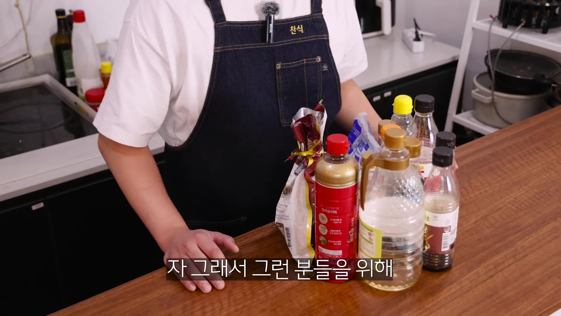 앞치마를 두른 요리사가 주방 조리대 위에 다양한 소스병과 식재료들을 올려두고 설명하는 모습