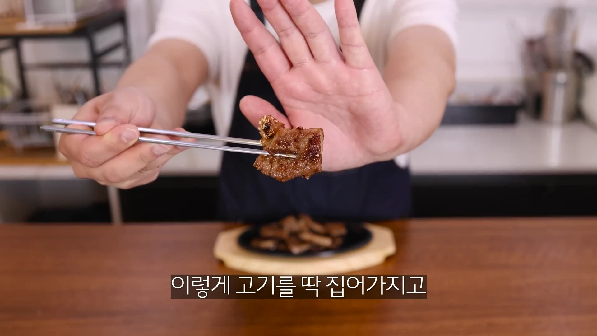 요리사가 한 손에는 젓가락으로 노릇하게 구워진 고기 한 점을 들고, 다른 한 손으로는 정면을 향해 손바닥을 펴 보이고 있다.