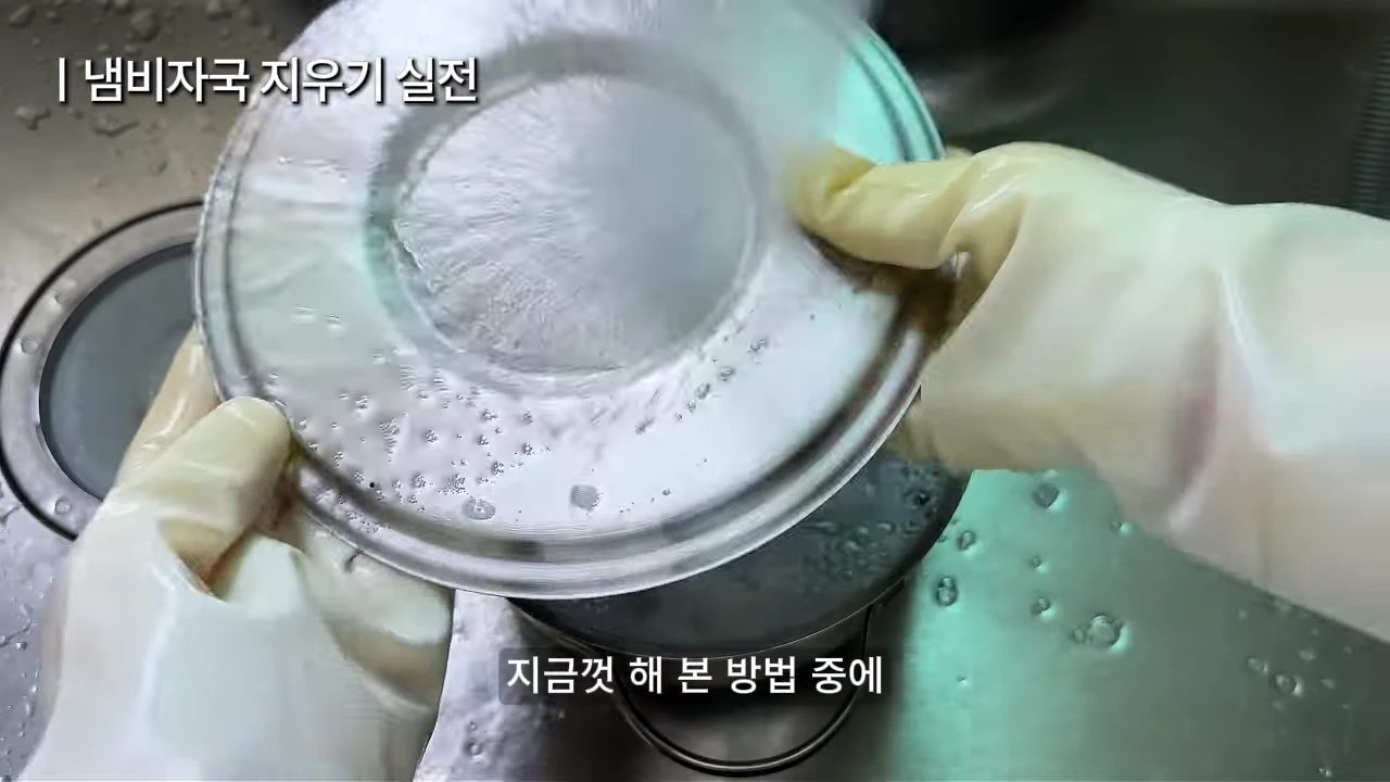 고무장갑을 낀 두 손이 스테인리스 냄비 뚜껑을 들고 세척하고 있는 모습이 담긴 영상 캡처 화면이며 상단에는 '냄비자국 지우기 실전', 하단에는 '지금껏 해 본 방법 중에'라는 자막이 표시되어 있다.