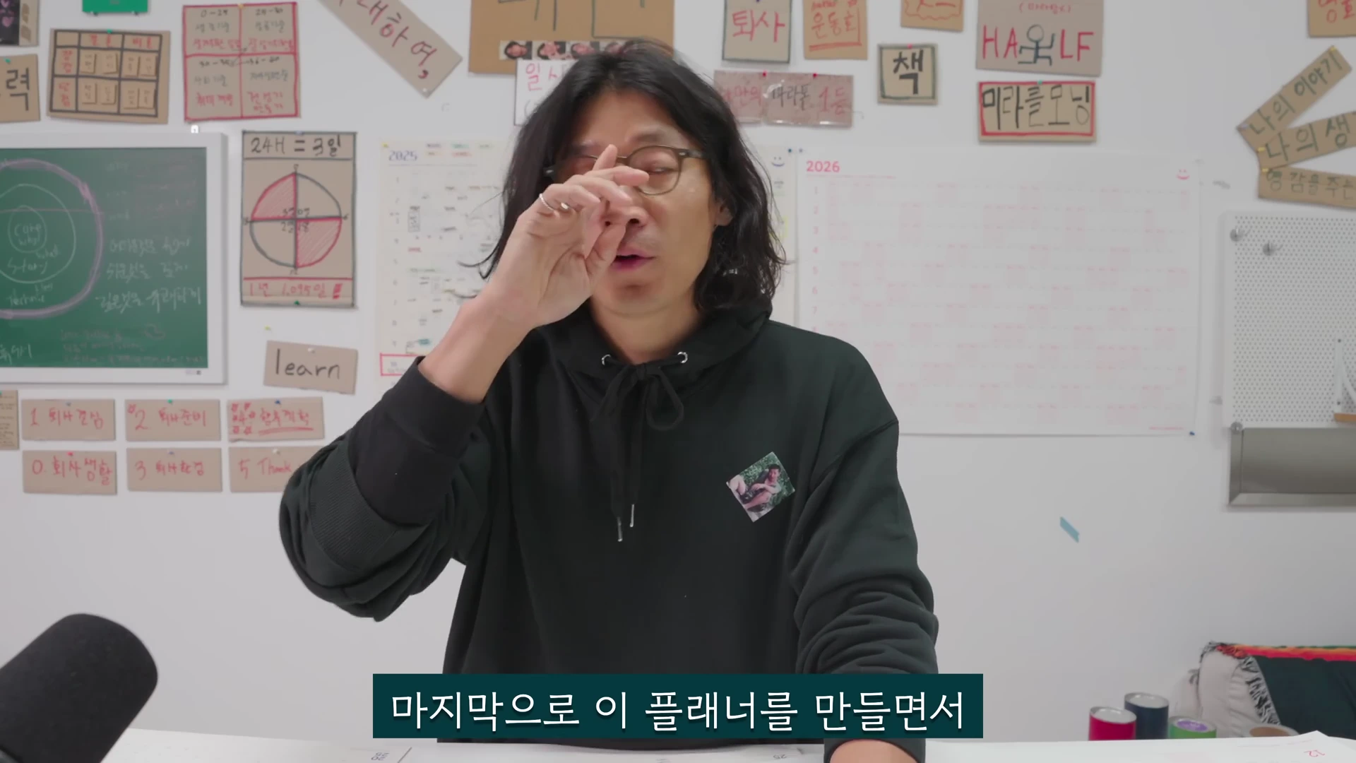 긴 머리에 안경을 쓴 남성이 검은색 후드티를 입고 손을 얼굴 근처에 올린 채 무언가 설명하고 있으며, 뒤쪽 벽면에는 다양한 문구와 그림이 붙은 게시판이 보입니다.