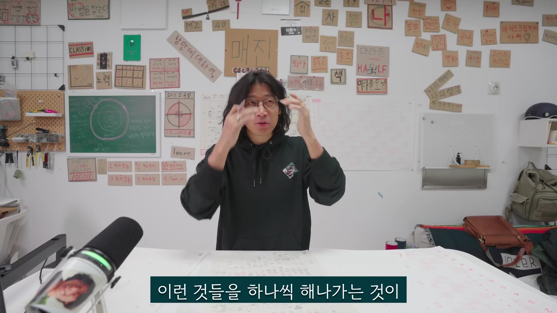 검은색 후드티를 입고 안경을 쓴 남성이 책상 앞에 앉아 양손을 머리 근처로 올린 채 설명하고 있으며, 뒤쪽 벽면에는 다양한 메모와 목표가 적힌 종이들이 붙어 있고 앞쪽에는 마이크와 플래너가 놓여 있다.