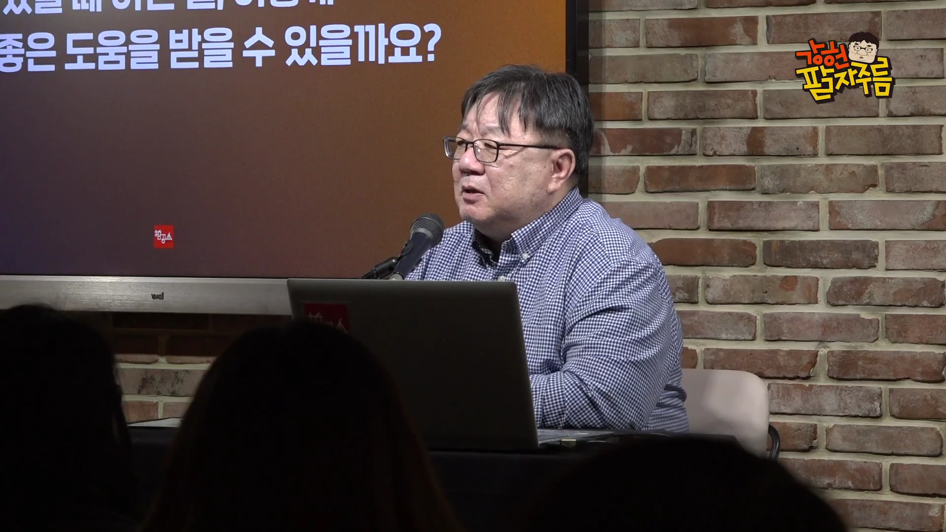안경을 쓴 중년 남성이 벽돌 벽 배경의 스튜디오에서 노트북과 마이크를 앞에 두고 앉아 청중에게 무언가를 이야기하고 있으며, 뒤쪽 스크린에는 한국어 문구가 일부 나타나 있습니다.