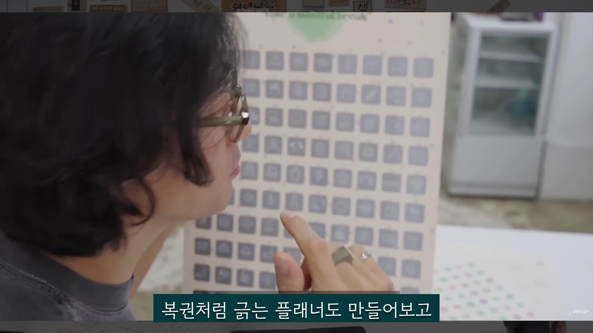 안경을 쓴 사람이 복권처럼 긁어 내용을 확인할 수 있는 격자무늬의 플래너 판을 손가락으로 가리키고 있다.